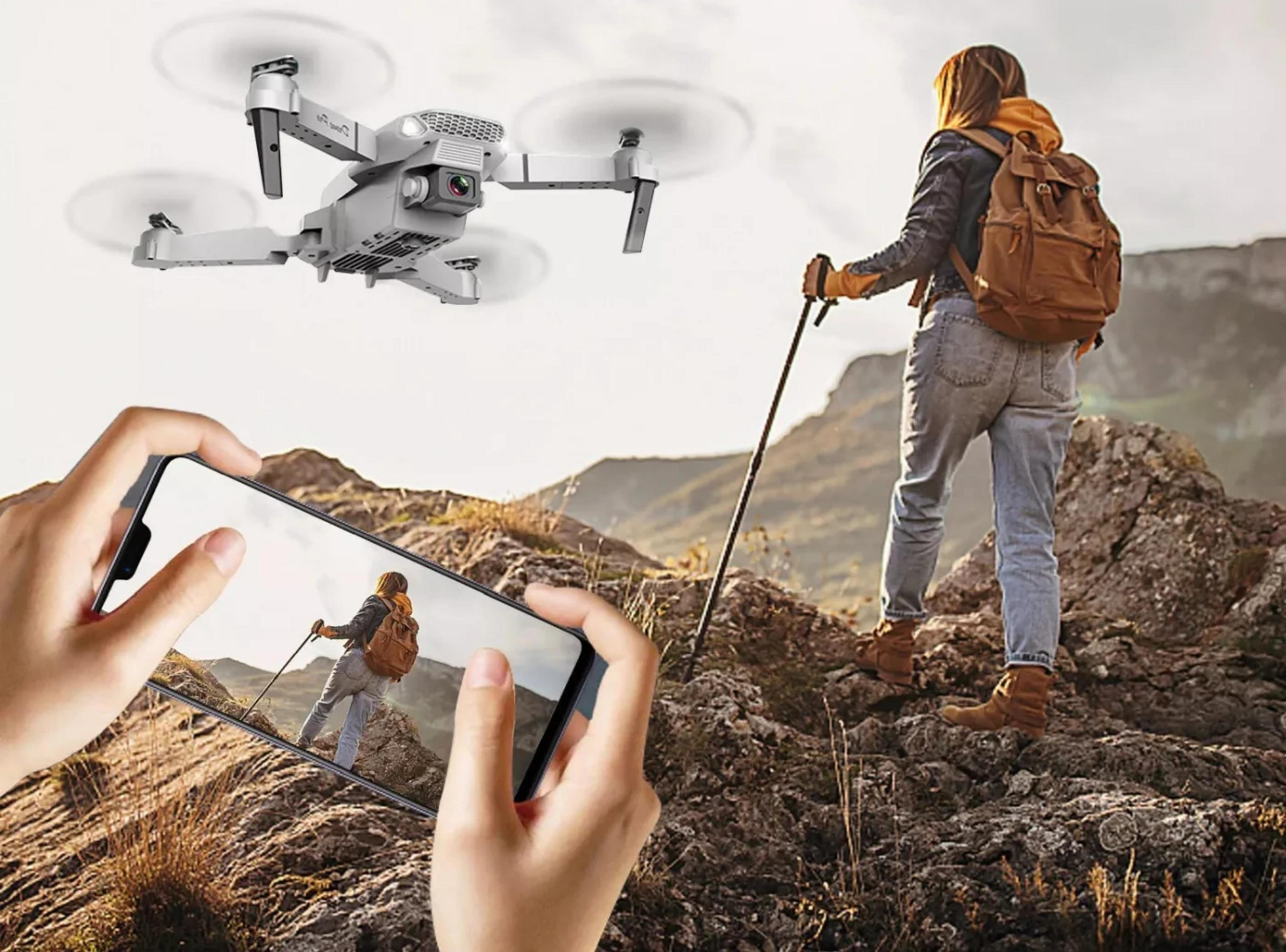 Un drone vola sopra un'escursionista con zaino e bastoni su terreno roccioso.