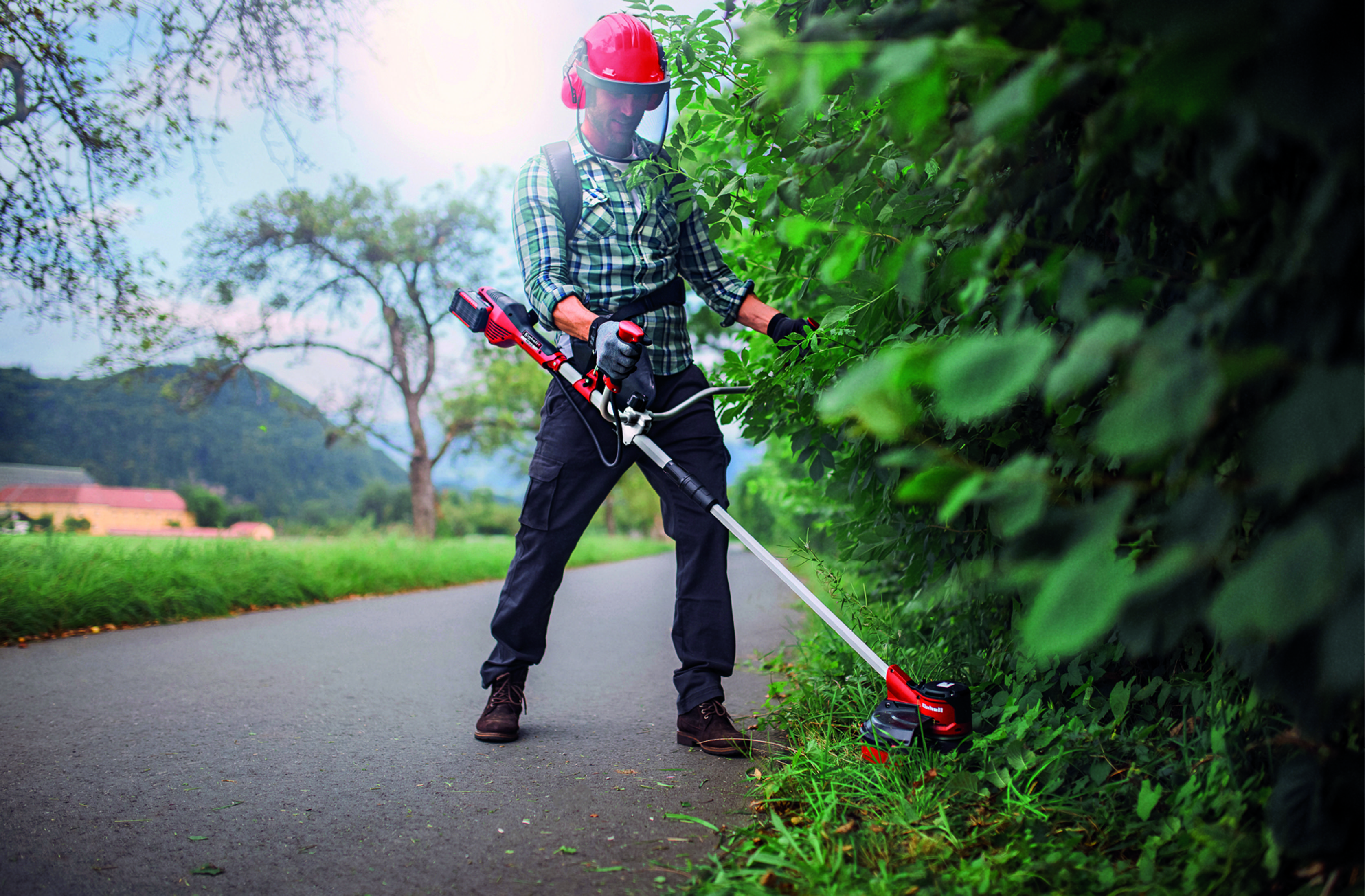 Ein Mann mit Helm trimmt Büsche mit einem Rasentrimmer auf einem Weg.