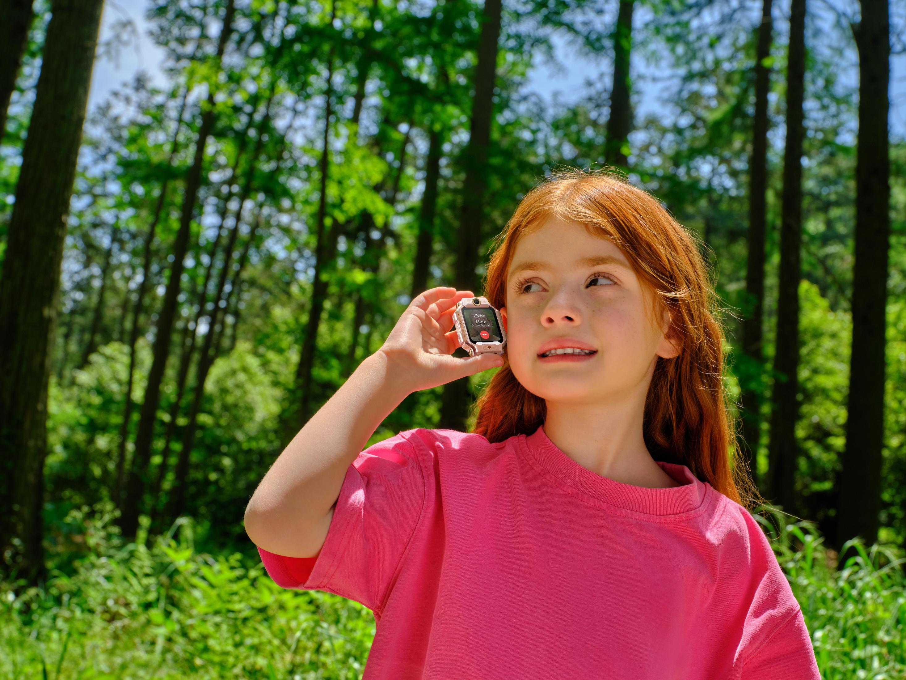 Ein rothaariges Mädchen mit einem pinken T-Shirt hält eine Smartwatch ans Ohr im Wald.