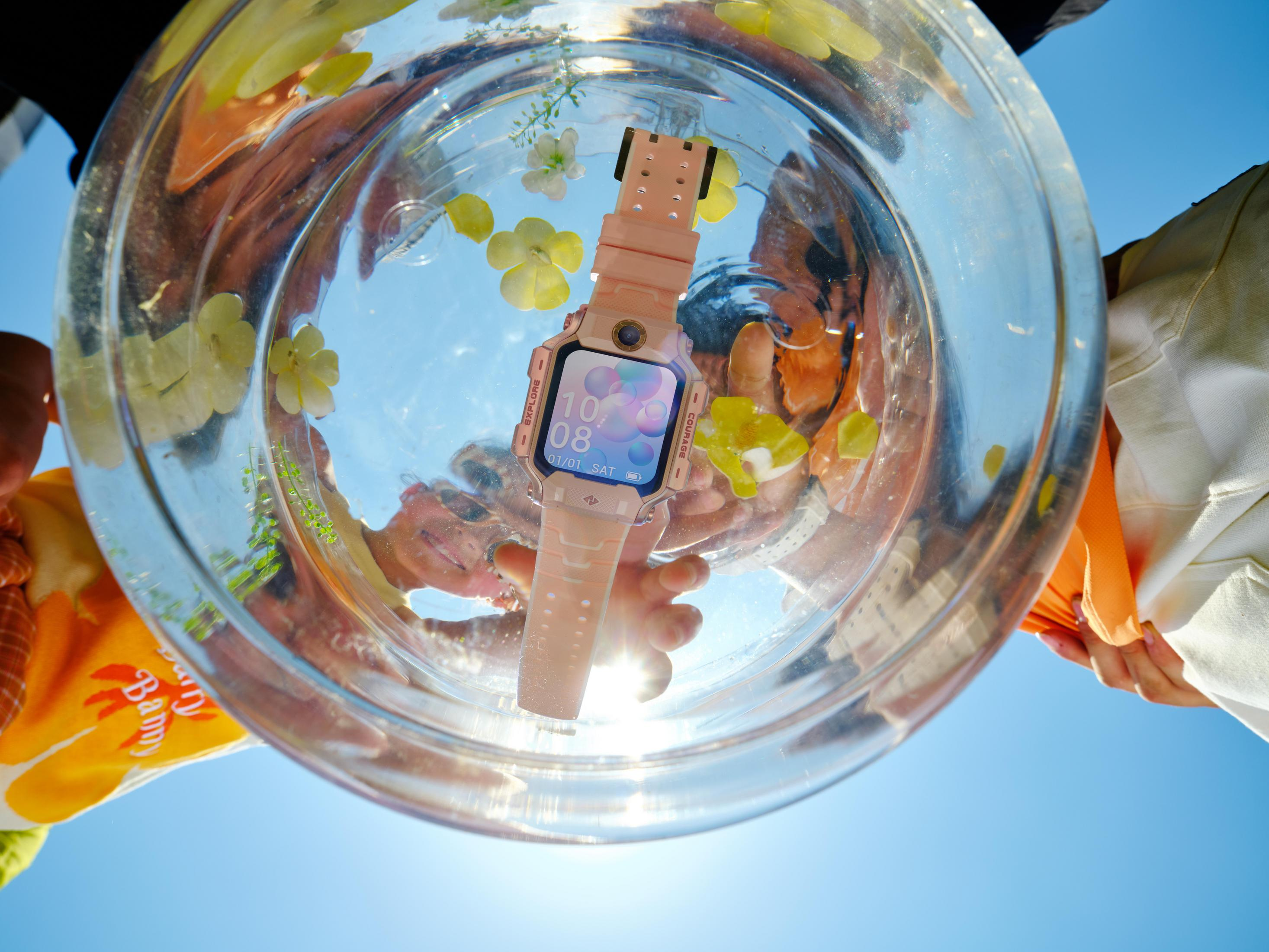 Eine rosa Smartwatch mit Blumen im Wasser, unter dem blauen Himmel gehalten.