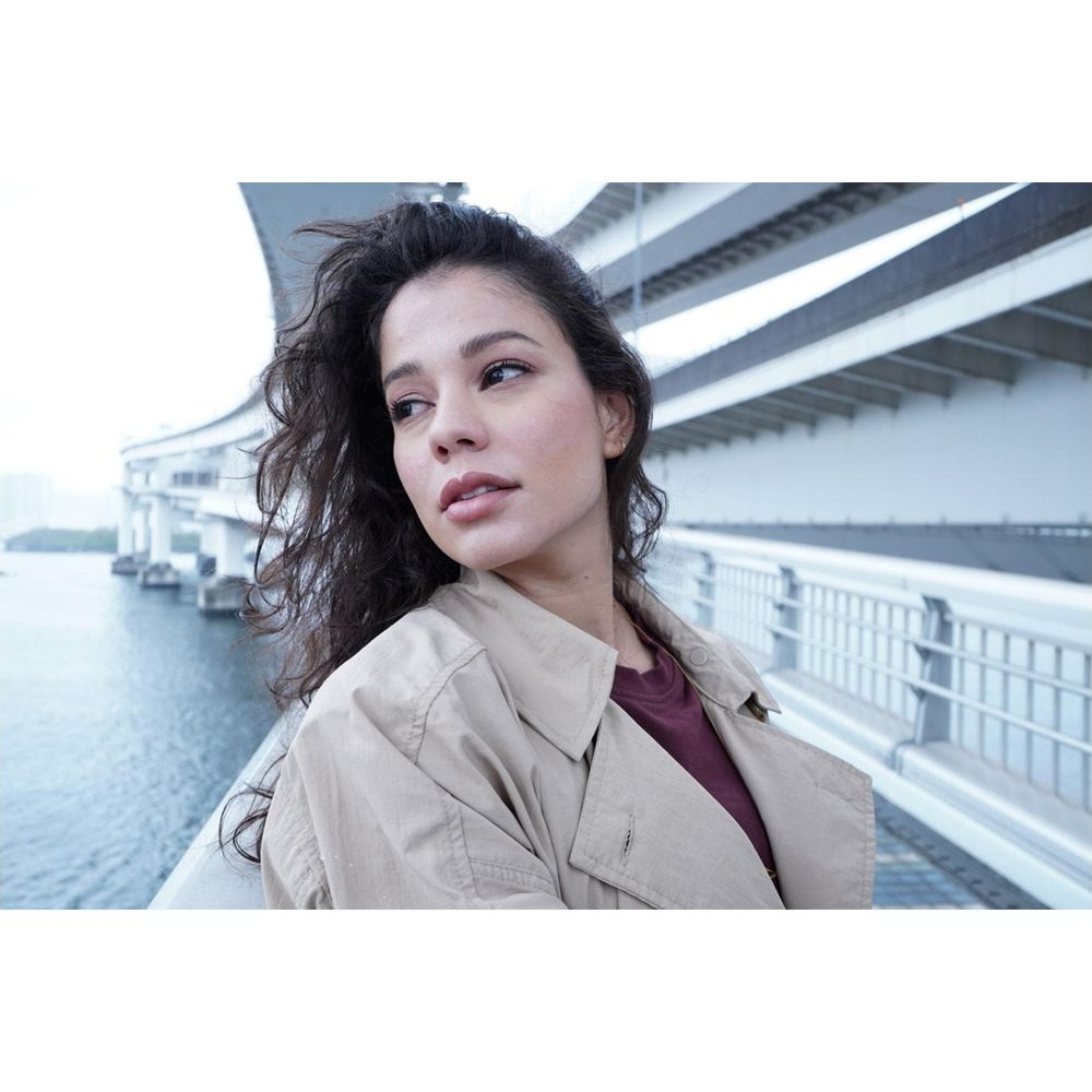 Vrouw met donker haar op een brug, met zee en reling op de achtergrond.