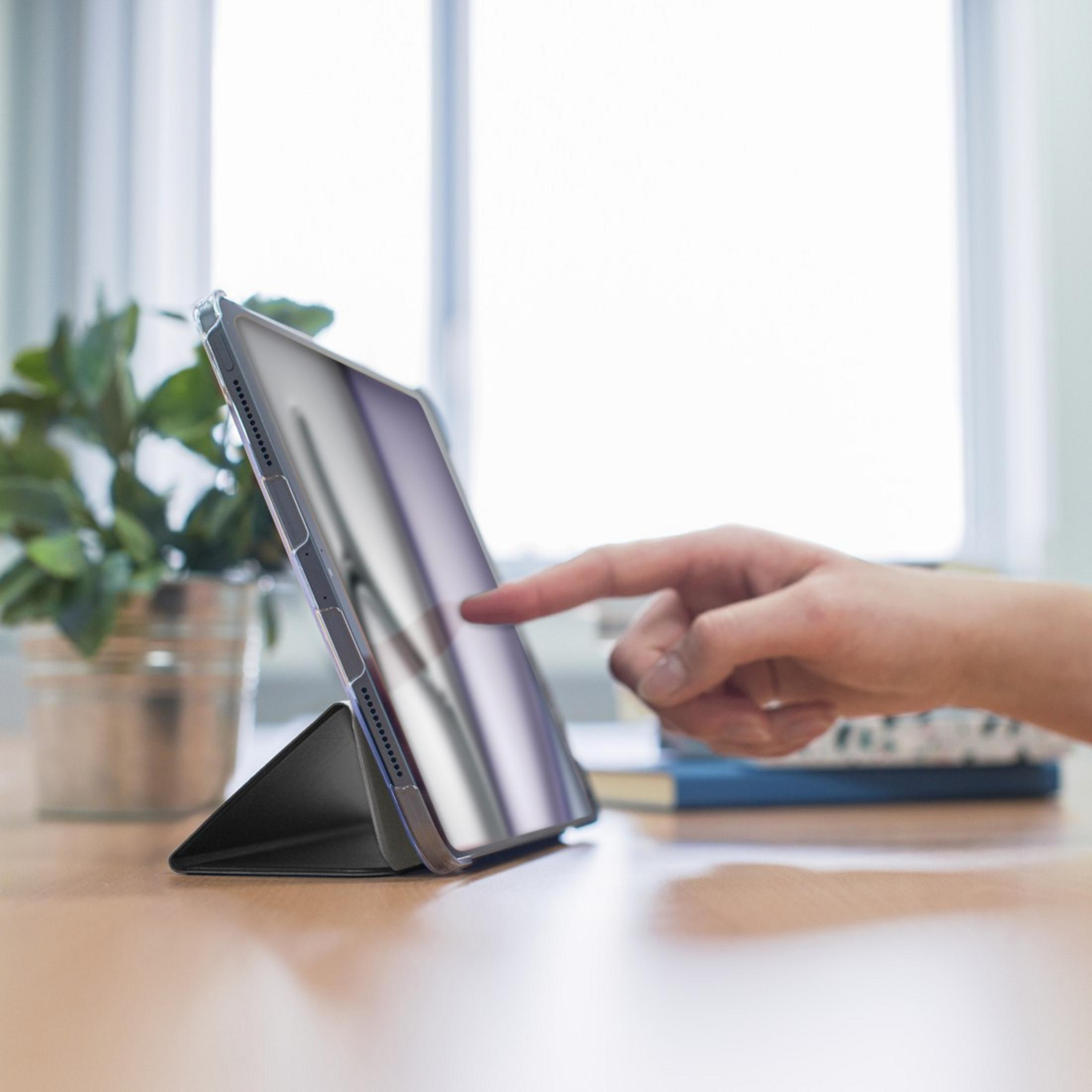 Eine Hand tippt auf ein Tablet, das auf einem Tisch steht. Daneben eine Pflanze.