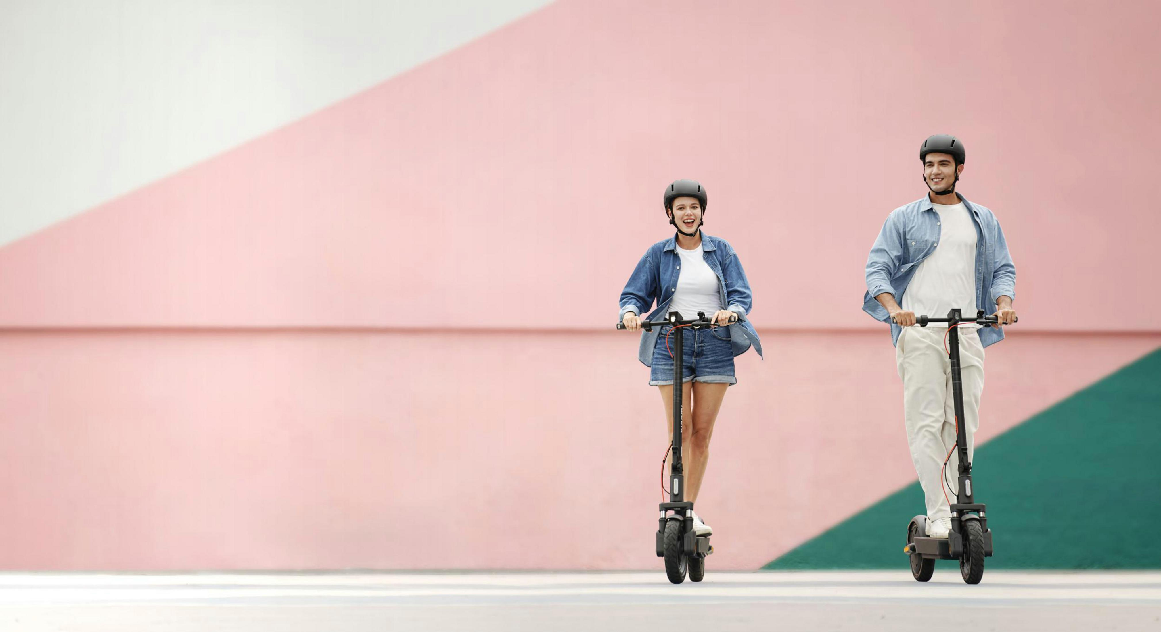Mann und Frau fahren E-Scooter vor einer rosa Wand.