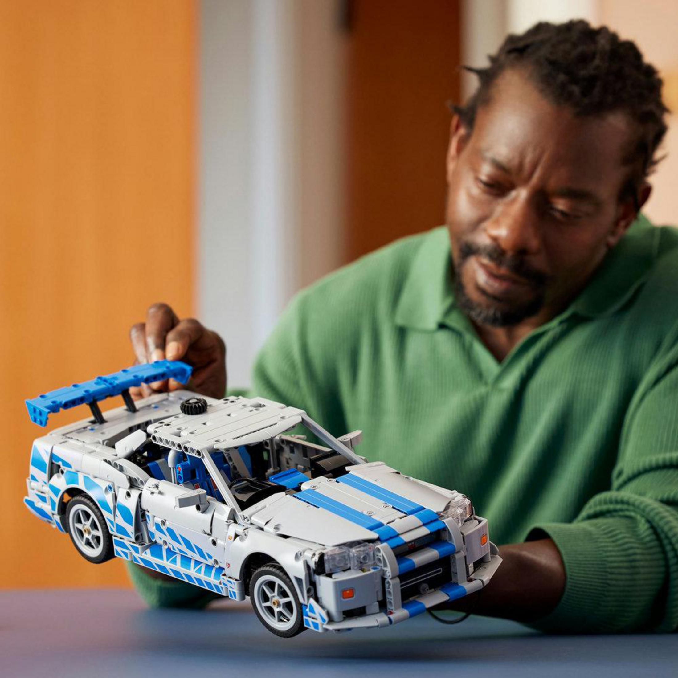Yeşil kazaklı adam, büyük kanatlı beyaz ve mavi Lego araba tutuyor.