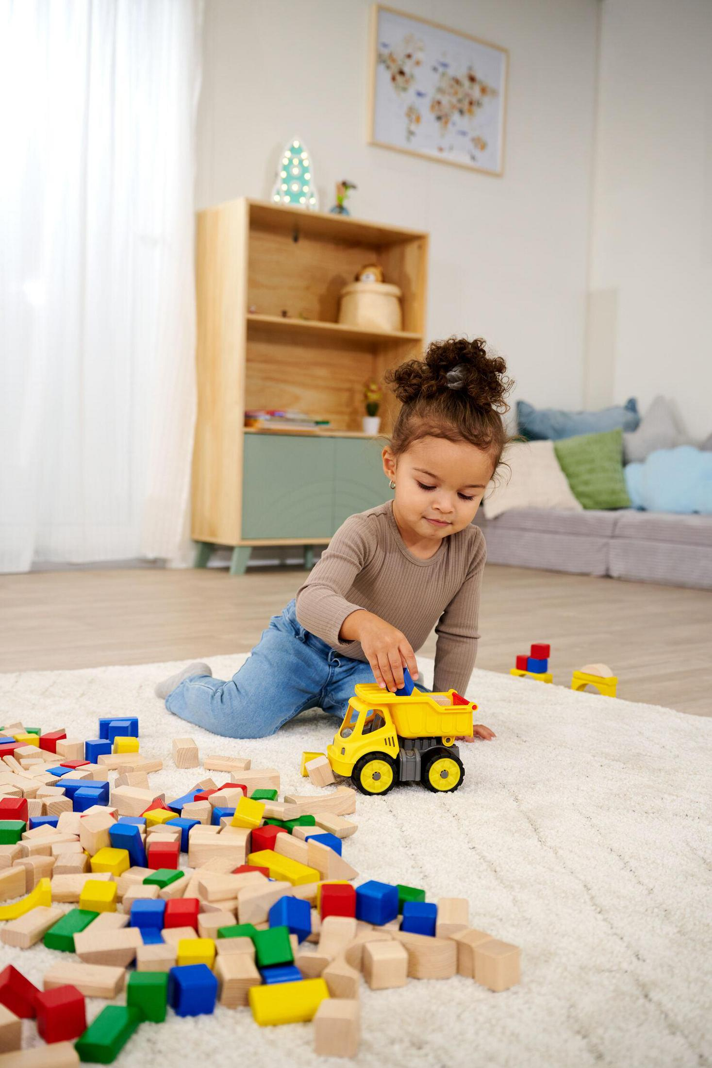 Una niña juega con bloques de madera coloridos y un camión de juguete amarillo sobre una alfombra.