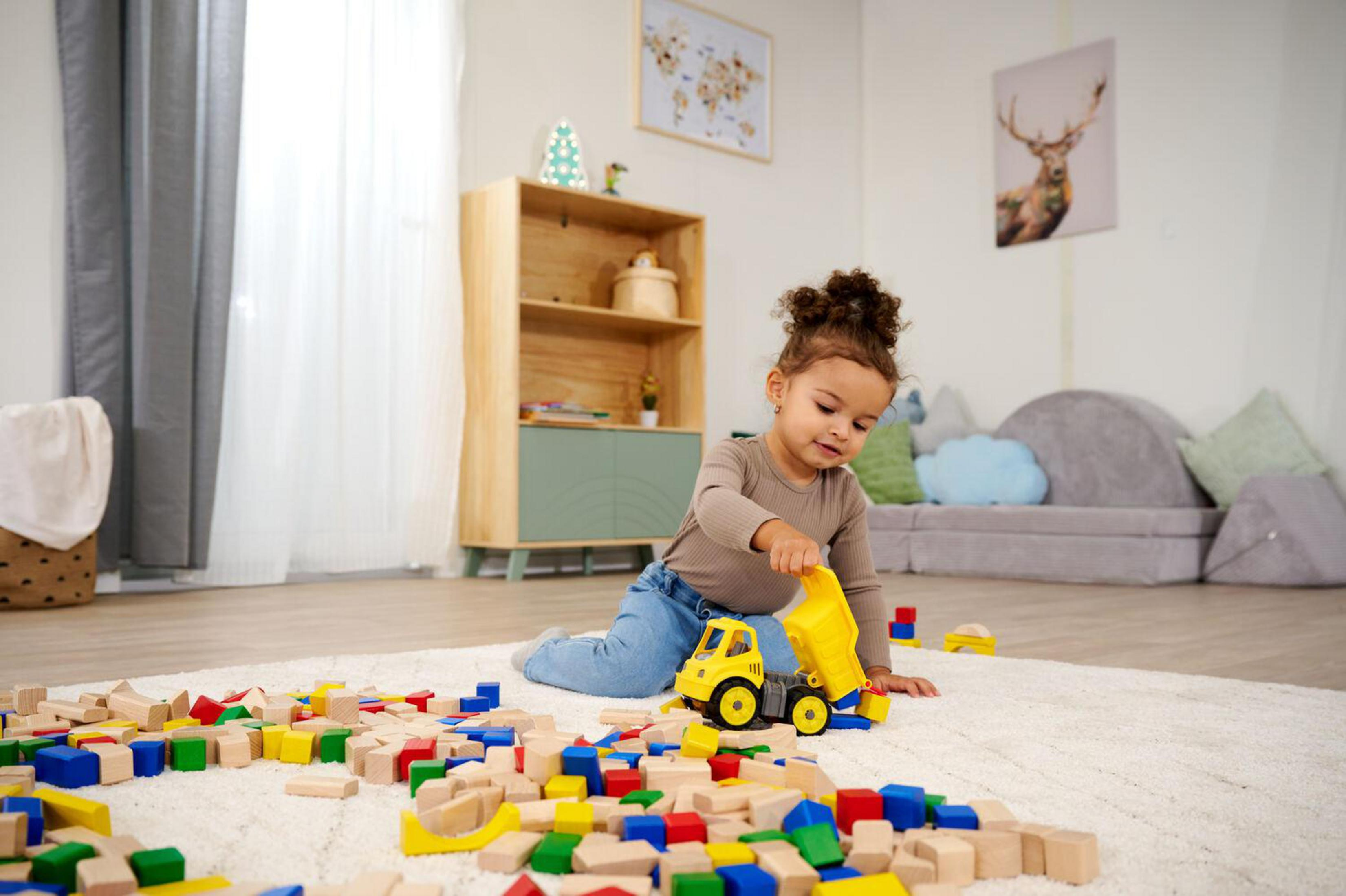 Una niña juega con bloques de construcción y un camión de juguete amarillo sobre una alfombra.