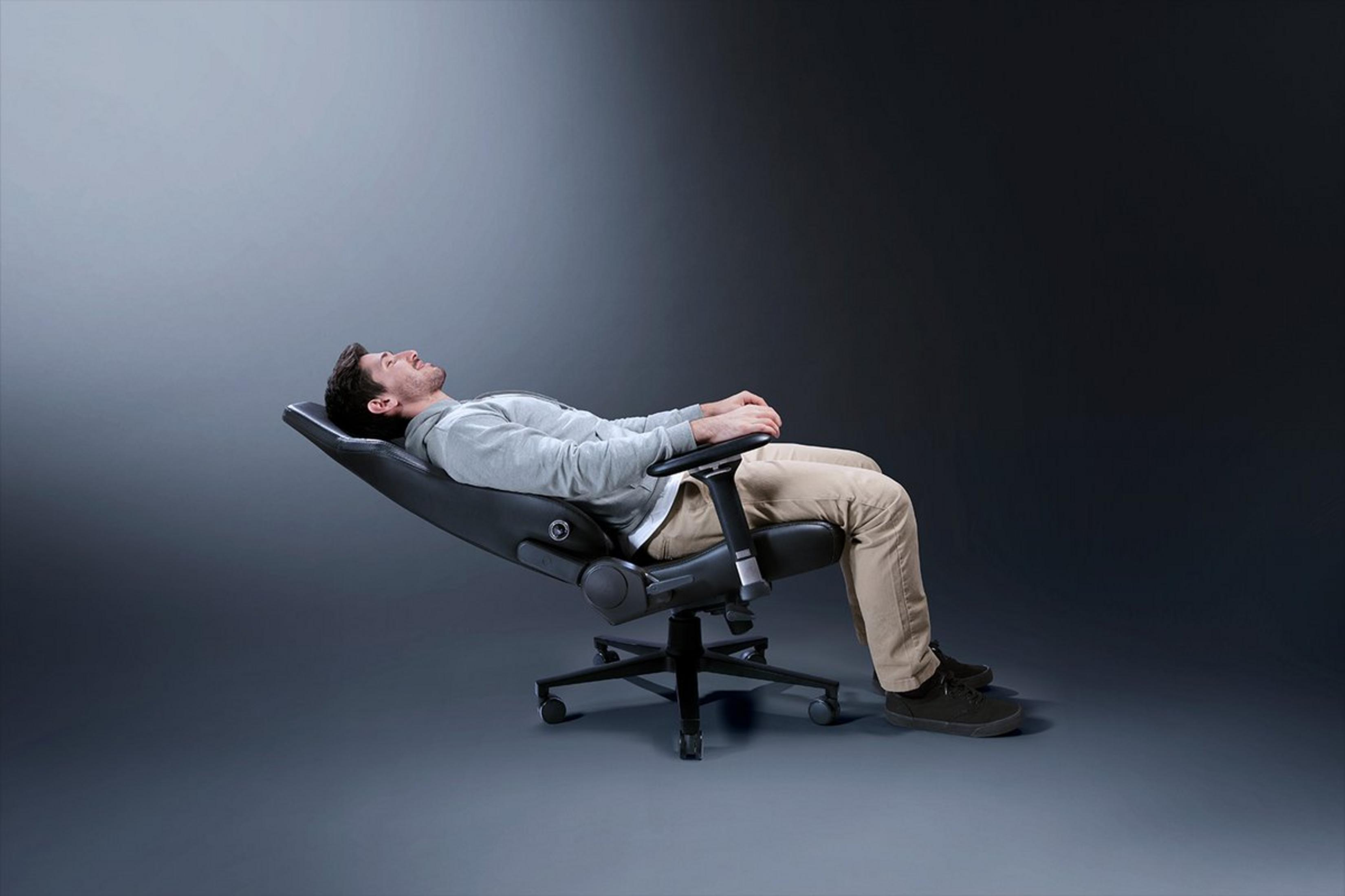 Uomo rilassato sdraiato su una sedia da ufficio nera, felpa grigia, pantaloni beige.