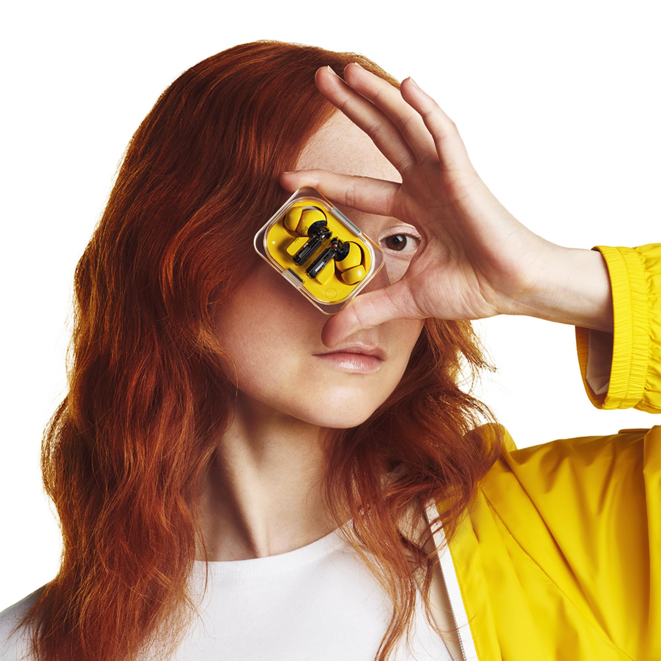 Vrouw met rood haar houdt gele oordopjes voor haar oog. Ze draagt een gele jas.