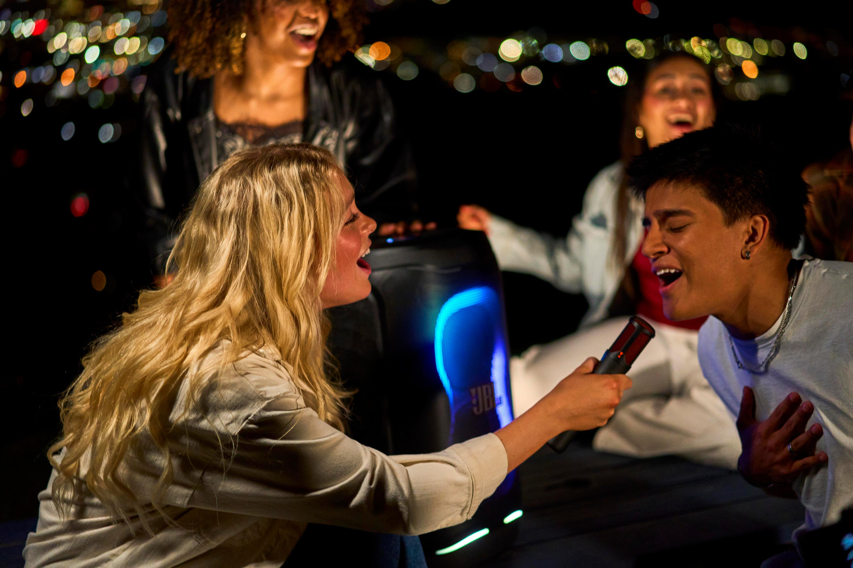 Gente canta en un micrófono junto a un altavoz brillante de noche con luces bokeh.