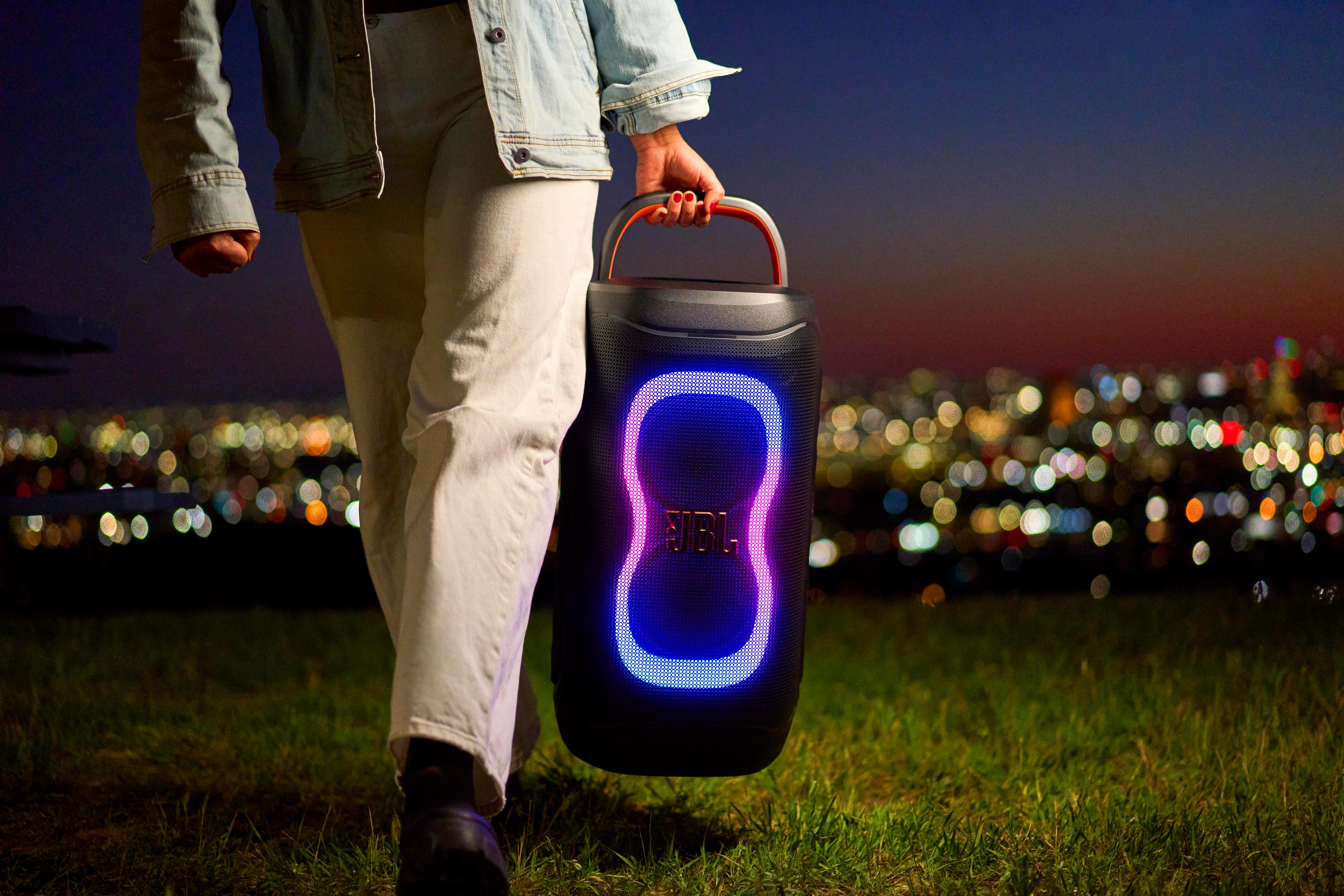 Una persona lleva un altavoz JBL iluminado al aire libre por la noche con luces de la ciudad al fondo.
