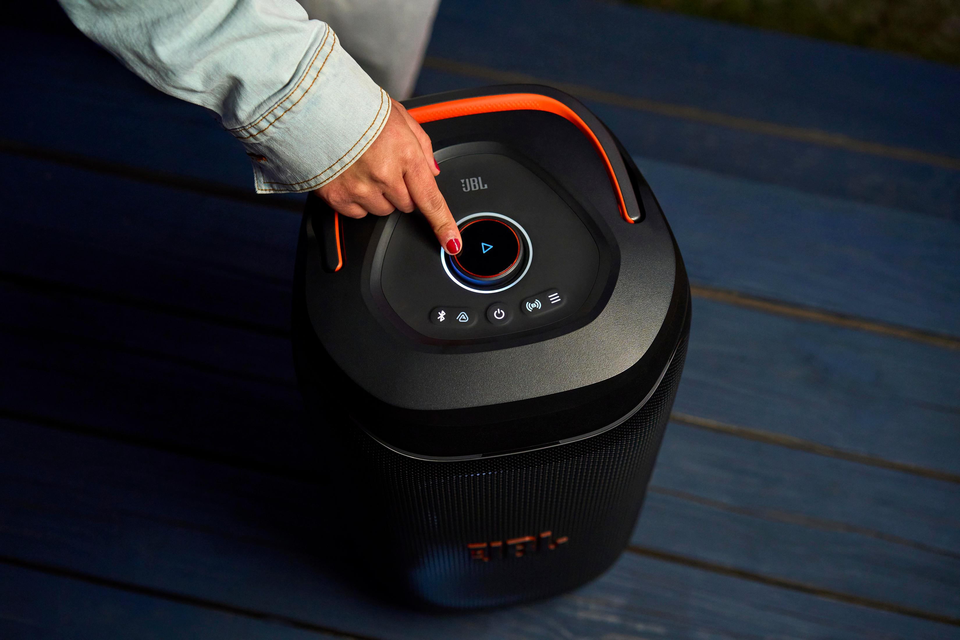 Una mano presiona el botón de reproducción en un altavoz JBL negro con detalles naranjas.