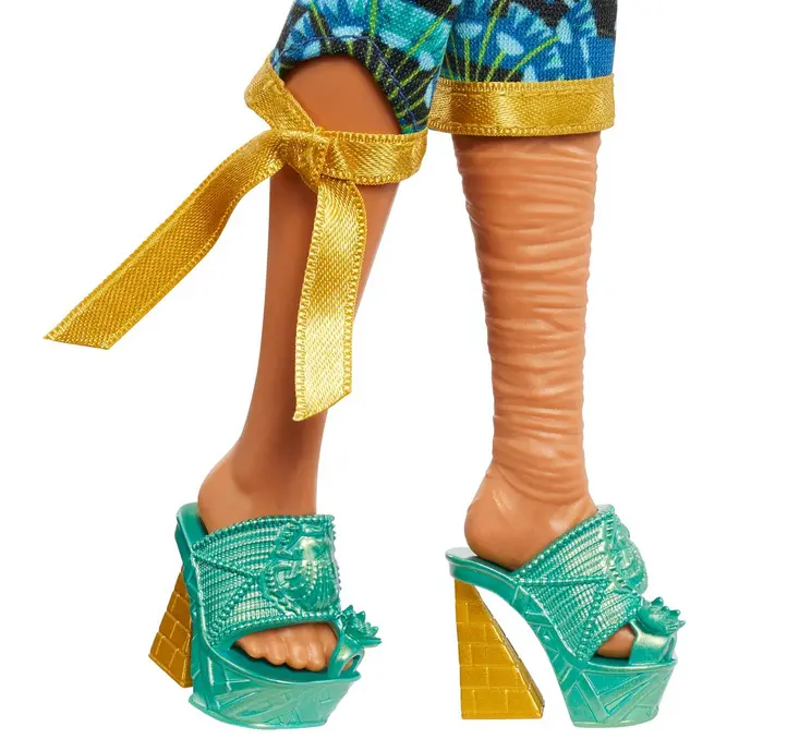 Eine Puppe trägt türkisfarbene Plateau-Sandalen mit goldenen Keilabsätzen und blau-grünen Hosen.