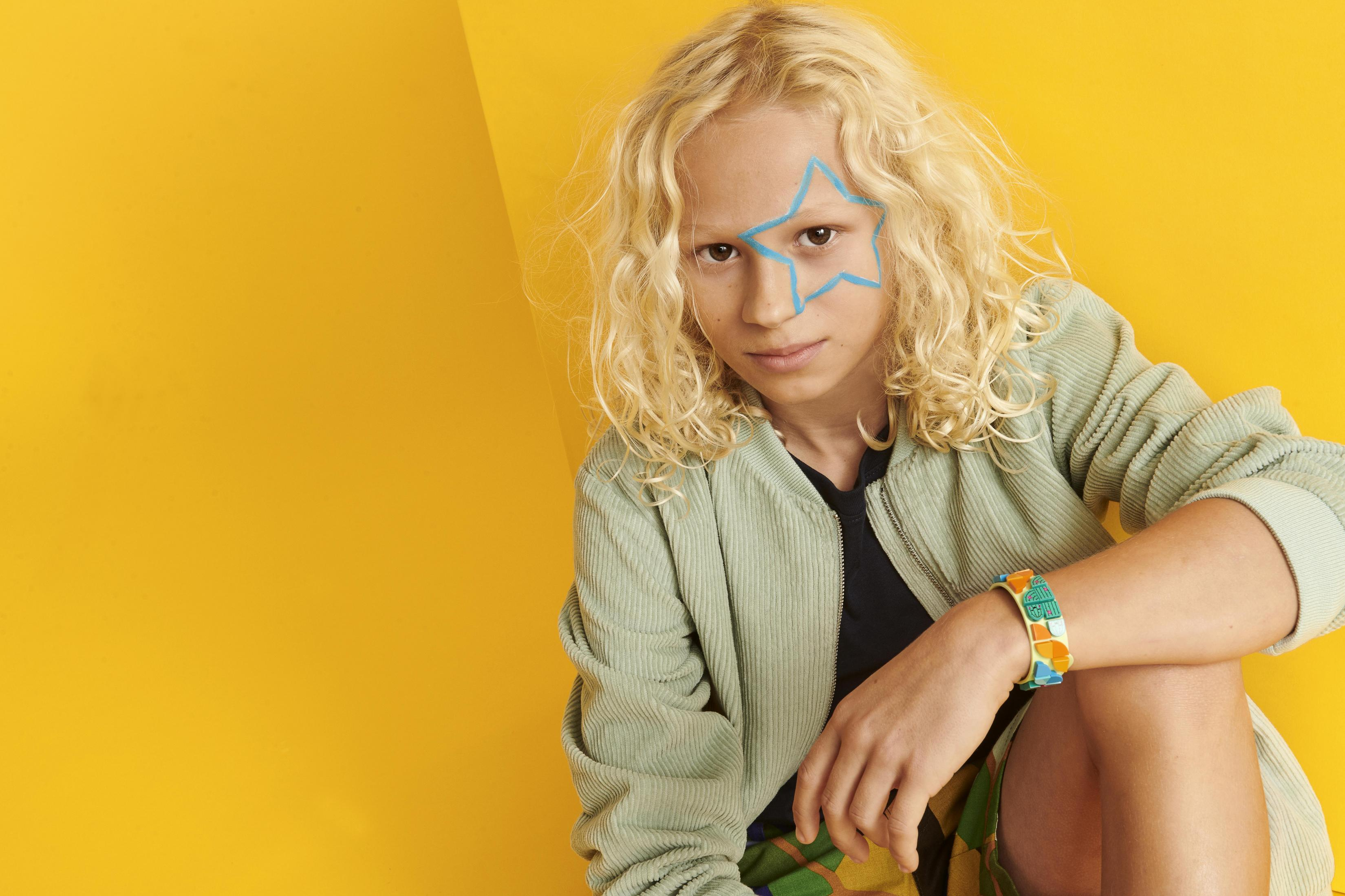 Un bambino con capelli ricci biondi e trucco a stella blu su sfondo giallo.