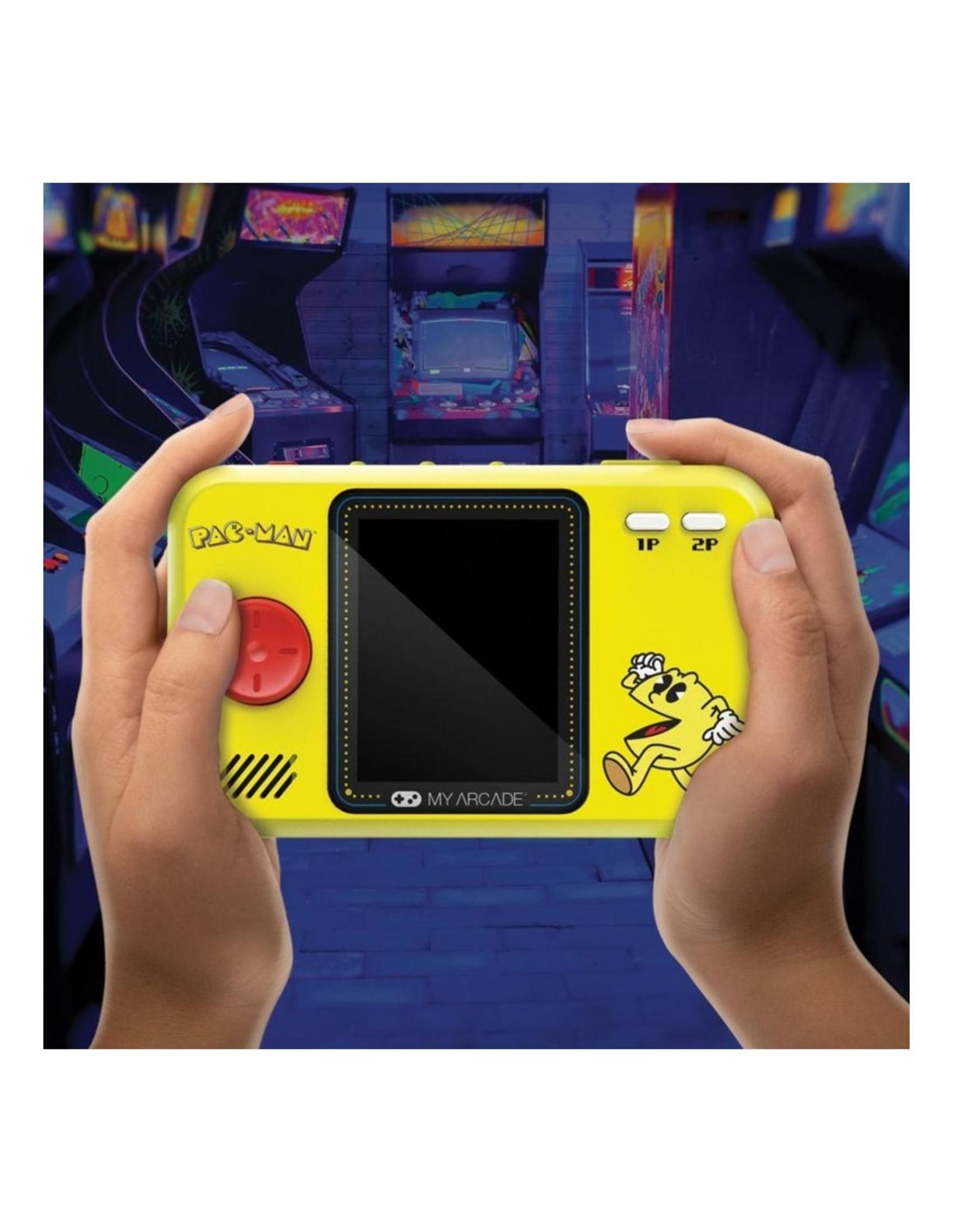 Manos sostienen un dispositivo de juego portátil amarillo de Pac-Man frente a gabinetes de arcade.