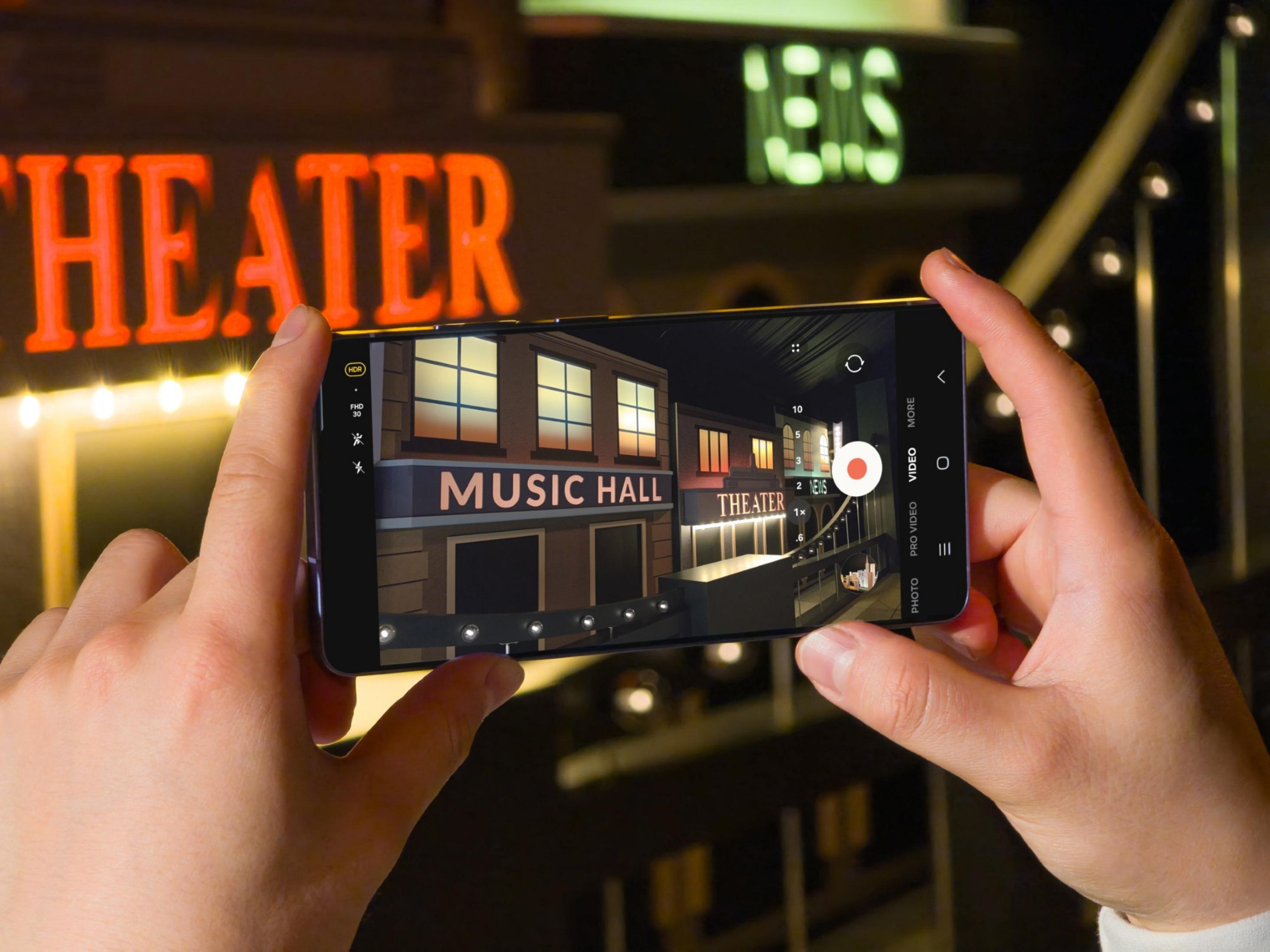 Eine Hand hält ein Smartphone, das eine beleuchtete Theaterfassade mit Schildern zeigt.