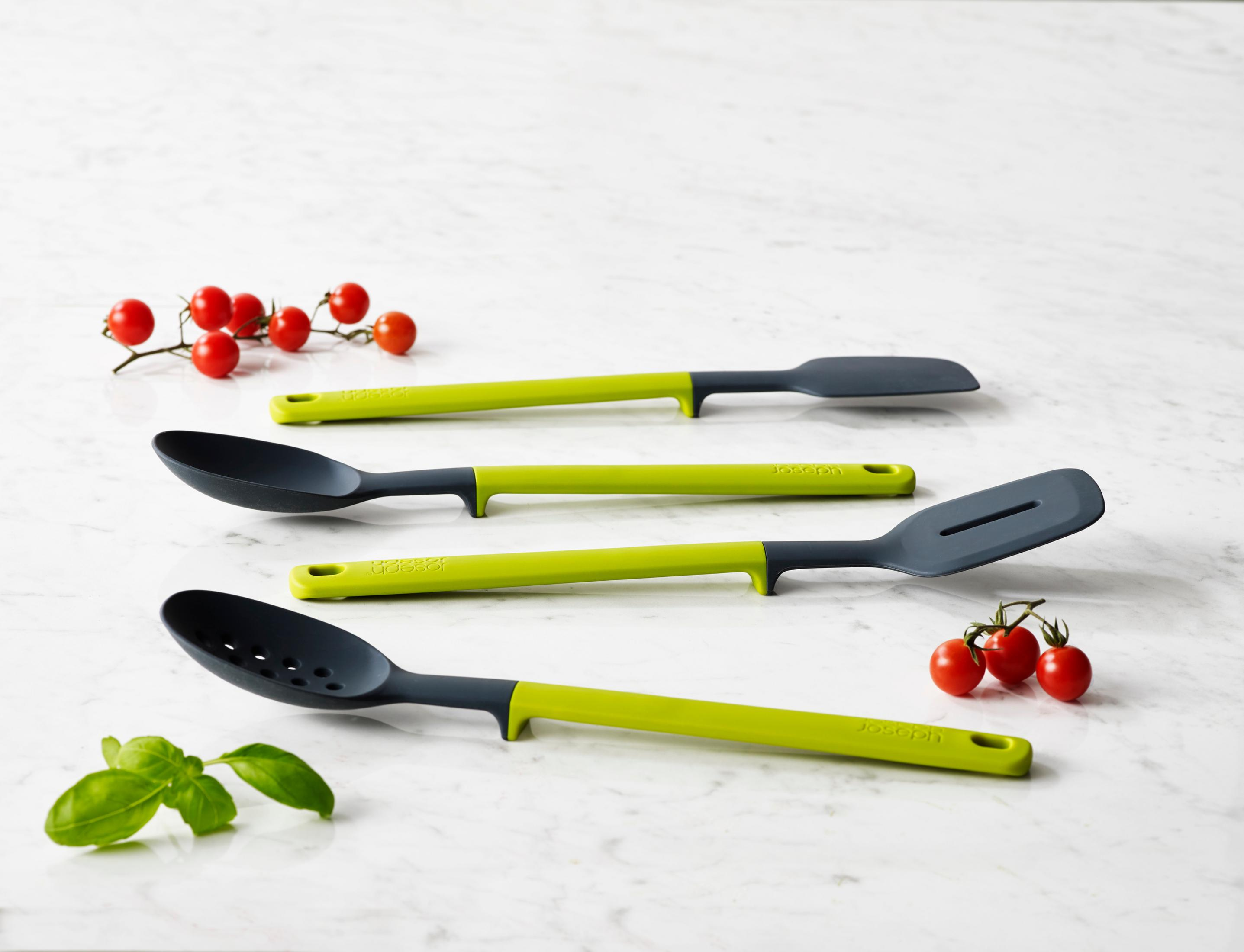 Cuatro utensilios de cocina con mangos verdes sobre superficie de mármol, con tomates y albahaca.