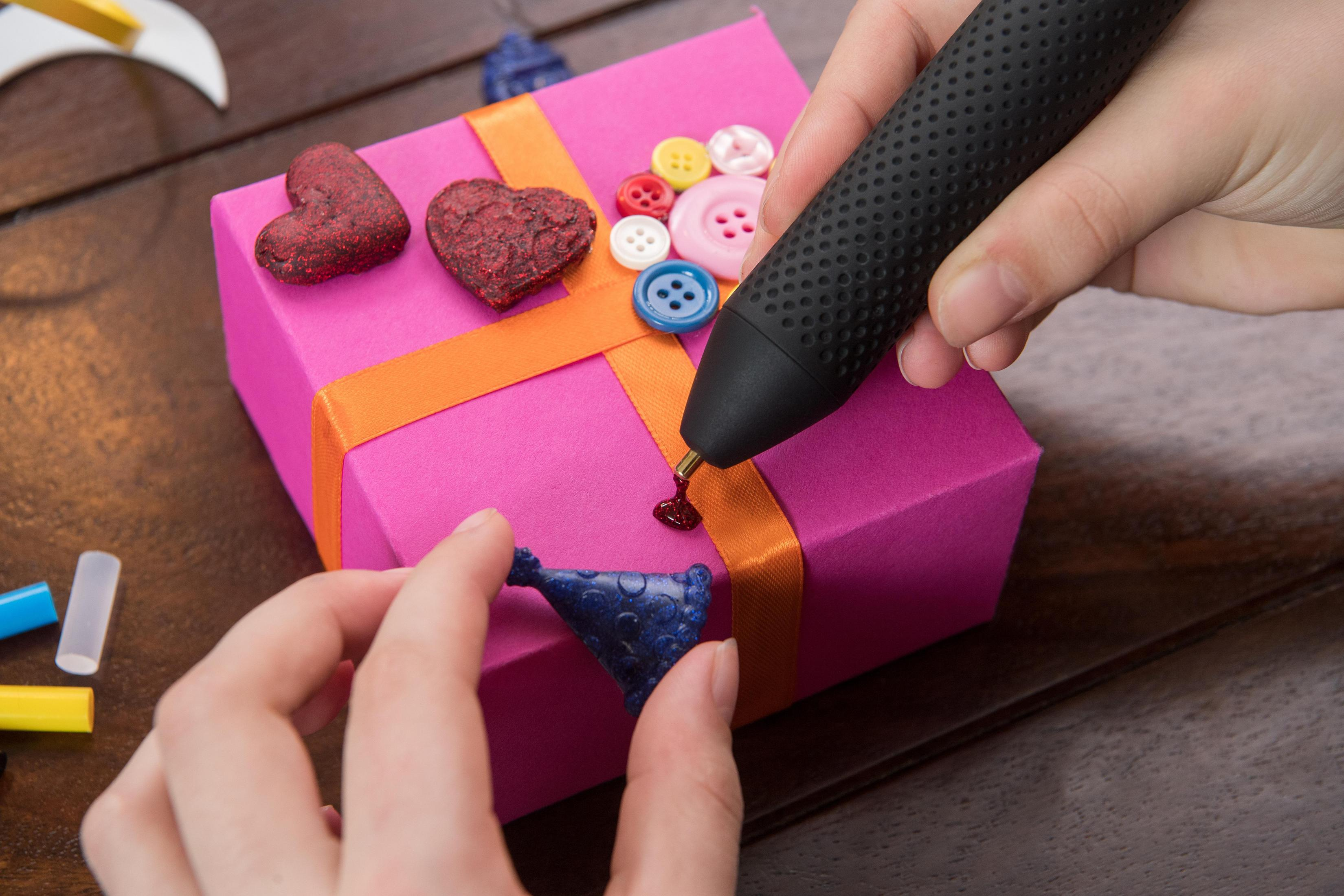Una mano sostiene un bolígrafo 3D negro que gotea sobre un regalo rosa con botones y corazones.