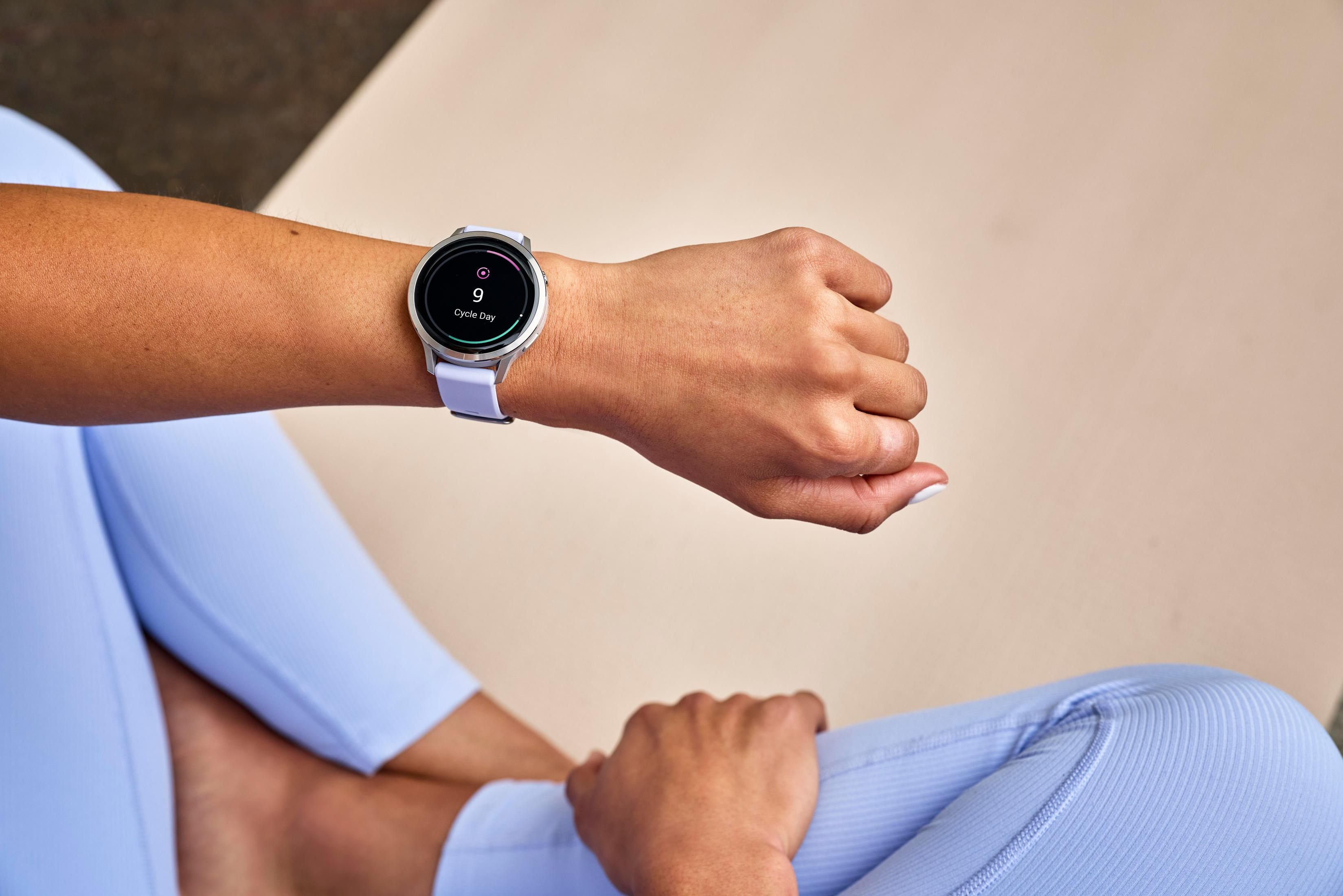 Das Handgelenk einer Person trägt eine silberne Smartwatch mit einem hellvioletten Band, auf dem 'Zyklustag 9' angezeigt wird.