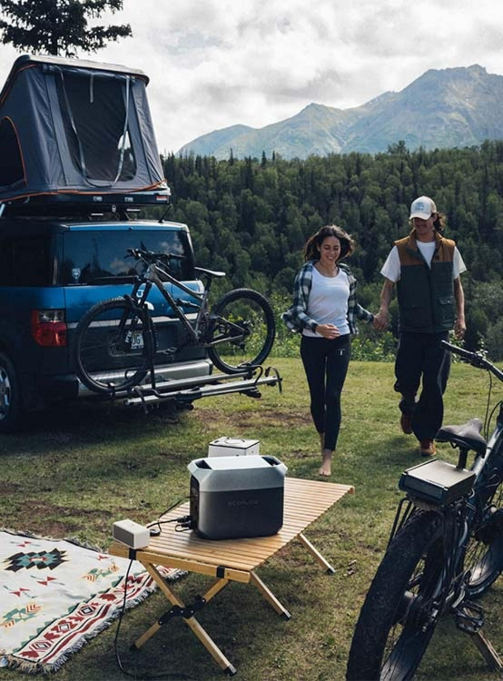 Ein Paar geht auf einem Campingplatz mit Dachzelt, Fahrrädern und Stromgenerator.