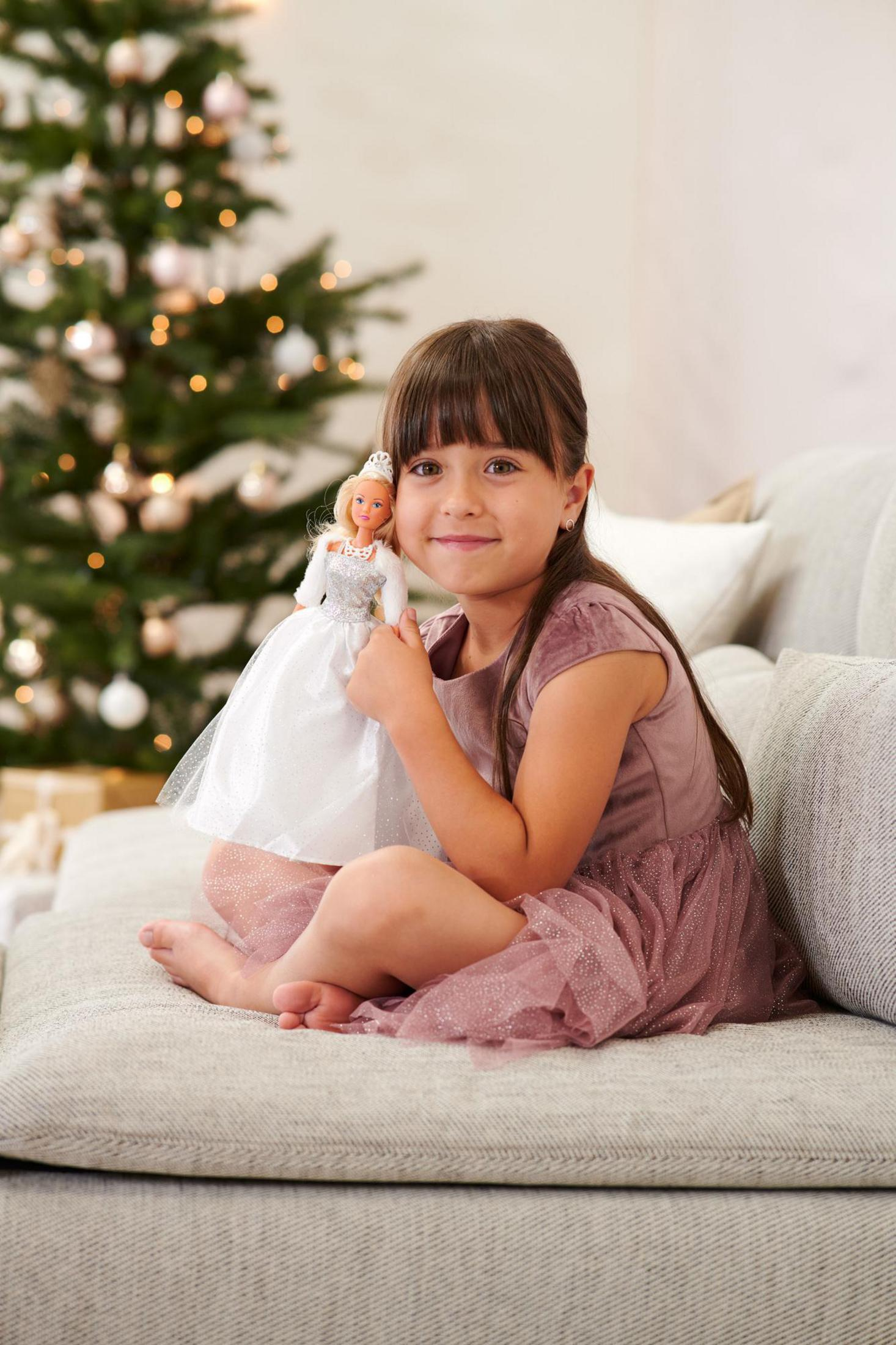 Ein junges Mädchen im rosa Kleid sitzt auf einer Couch und hält eine Puppe. Ein Weihnachtsbaum ist im Hintergrund.
