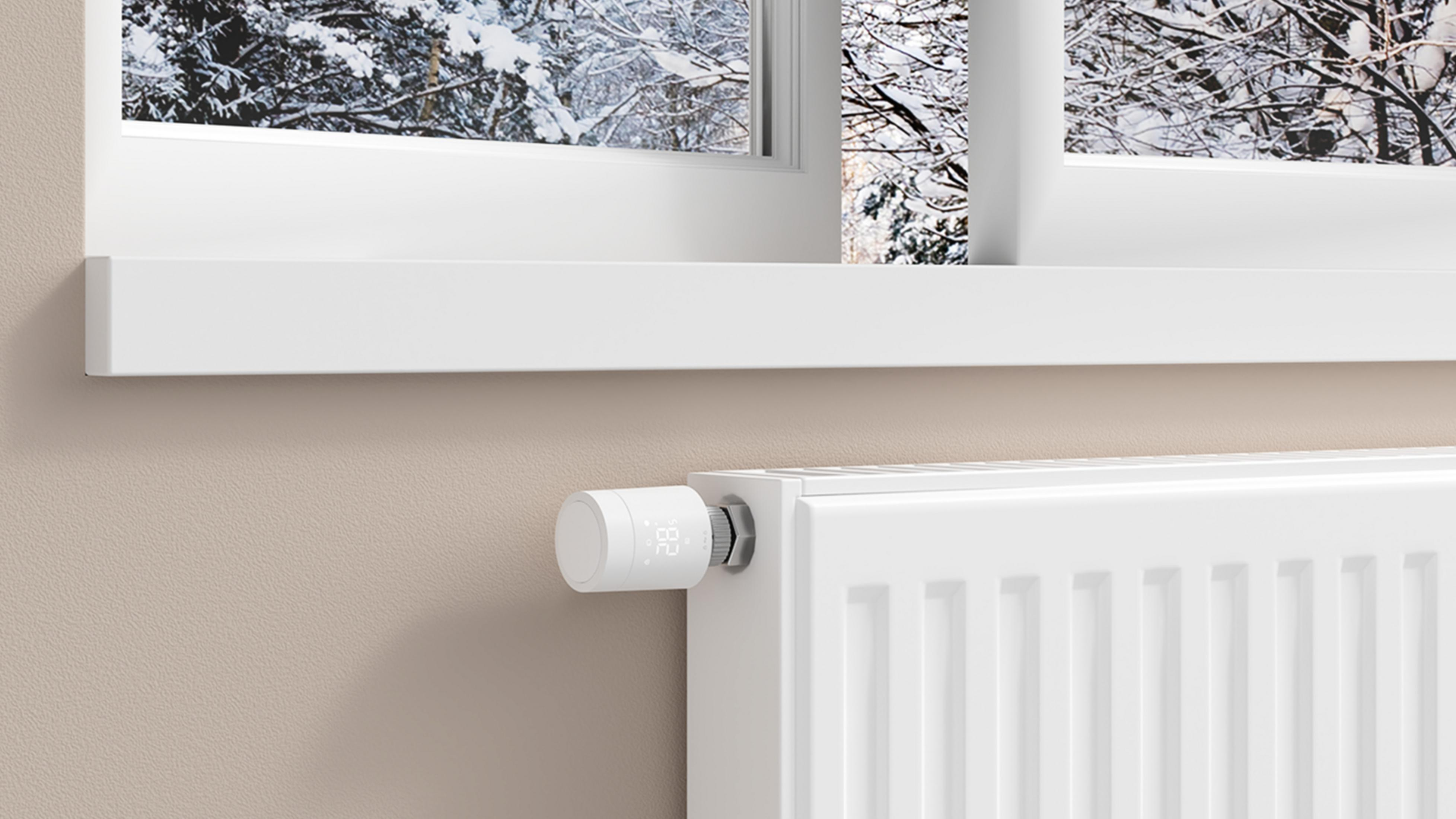 Ein weißer Heizkörperthermostat zeigt 28 Grad an, vor verschneiten Bäumen.