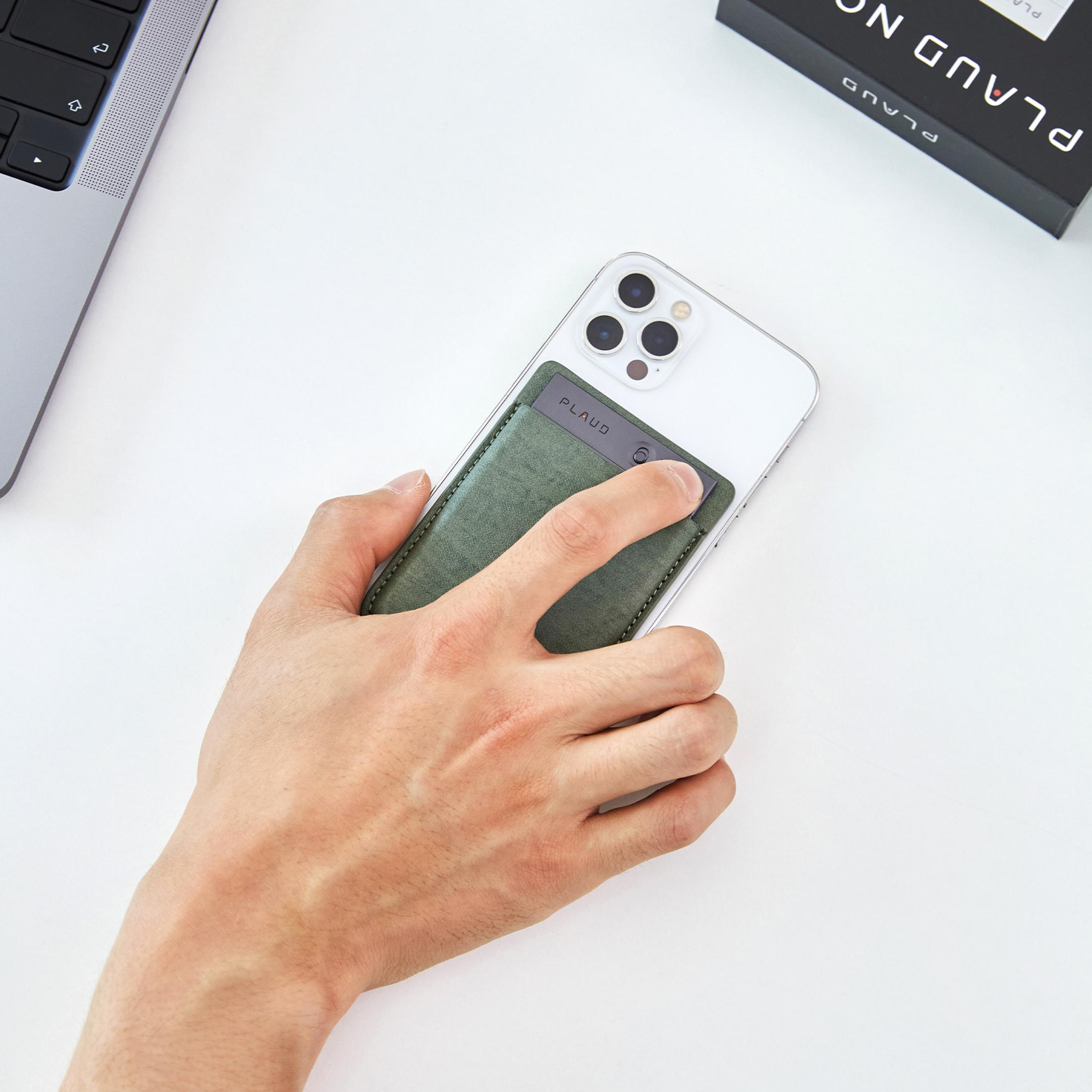 Eine Hand hält ein Handy mit einer grünen Tasche, daneben ein Laptop und eine Box.