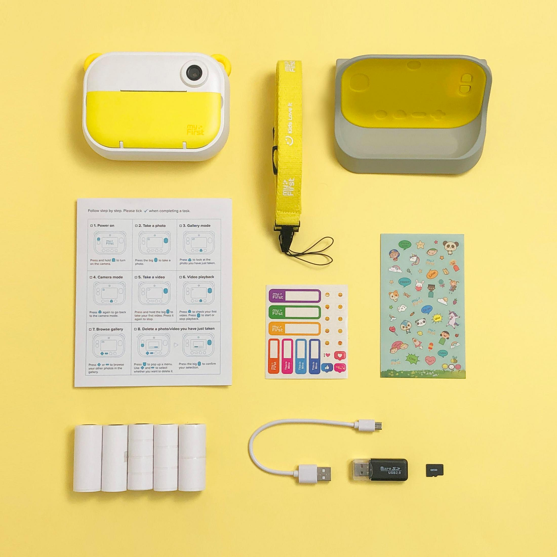 Ein gelbes und weißes Spielzeug-Kamera-Set mit Anleitung, Stickern, USB-Kabeln und Papierrollen.