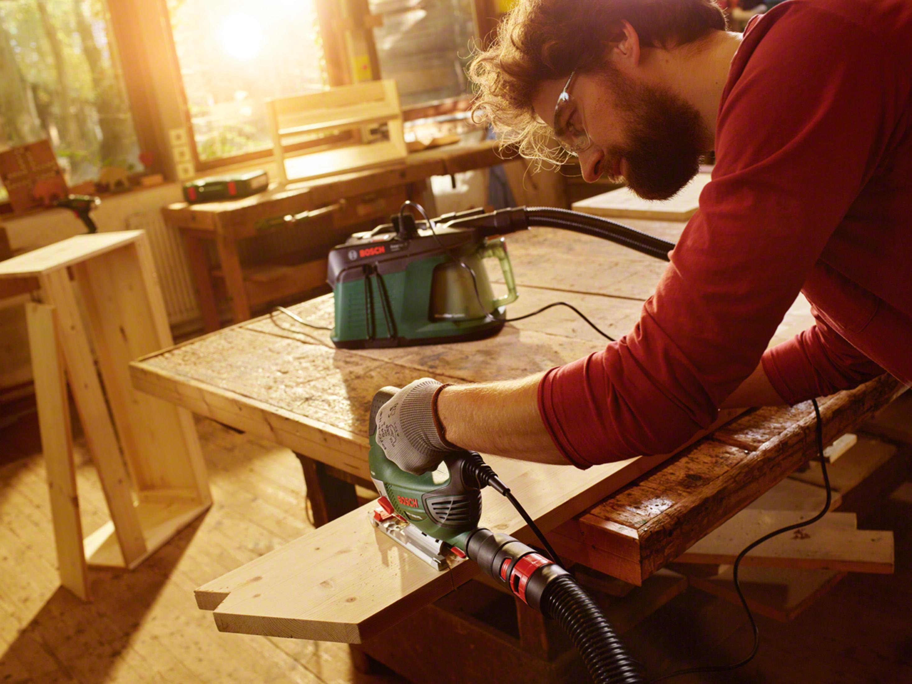 Un uomo con occhiali e barba taglia un asse di legno con un seghetto alternativo in un'officina.