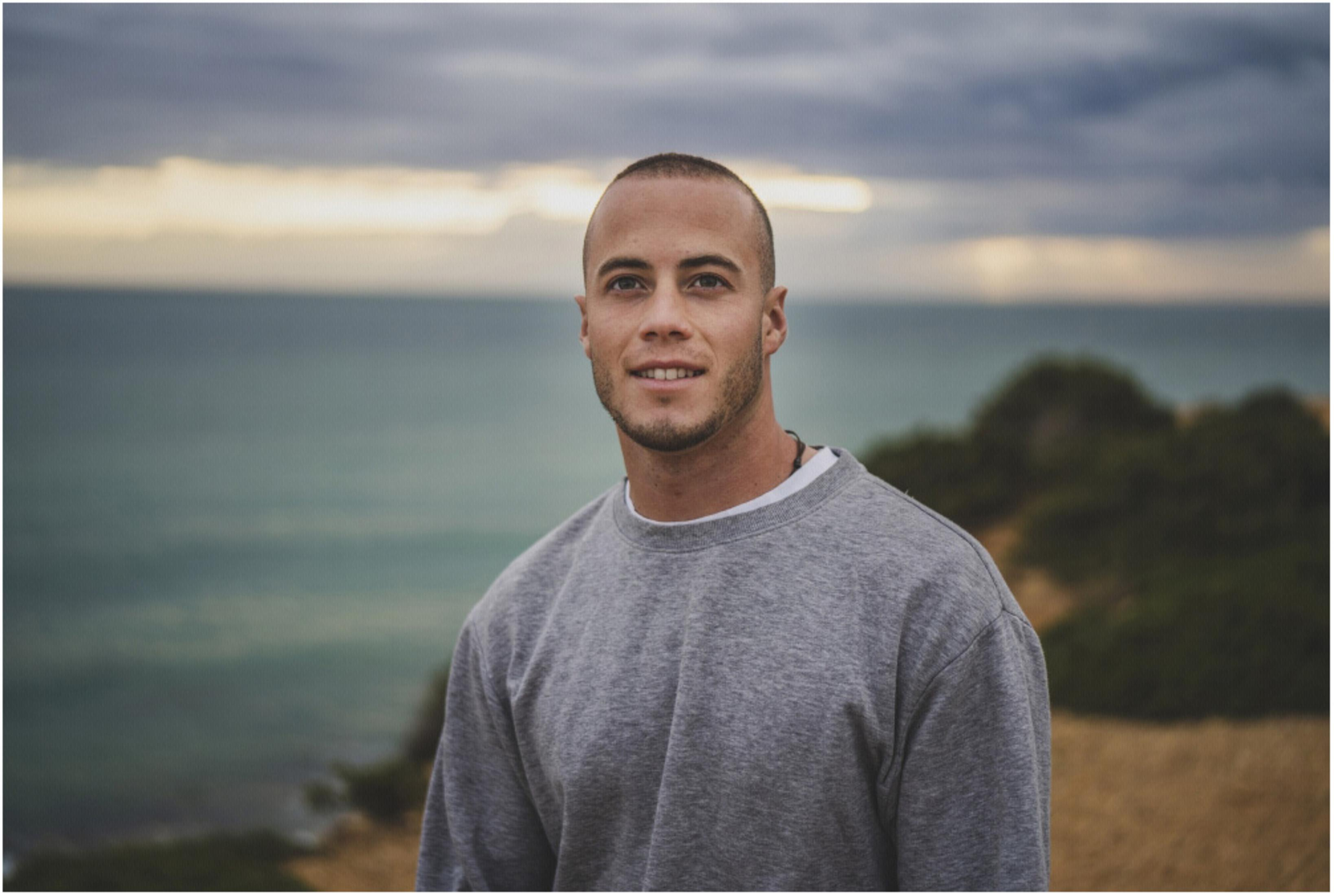 Mann mit grauem Sweatshirt und kurzem Haar blickt lächelnd in die Kamera, Meer im Hintergrund.