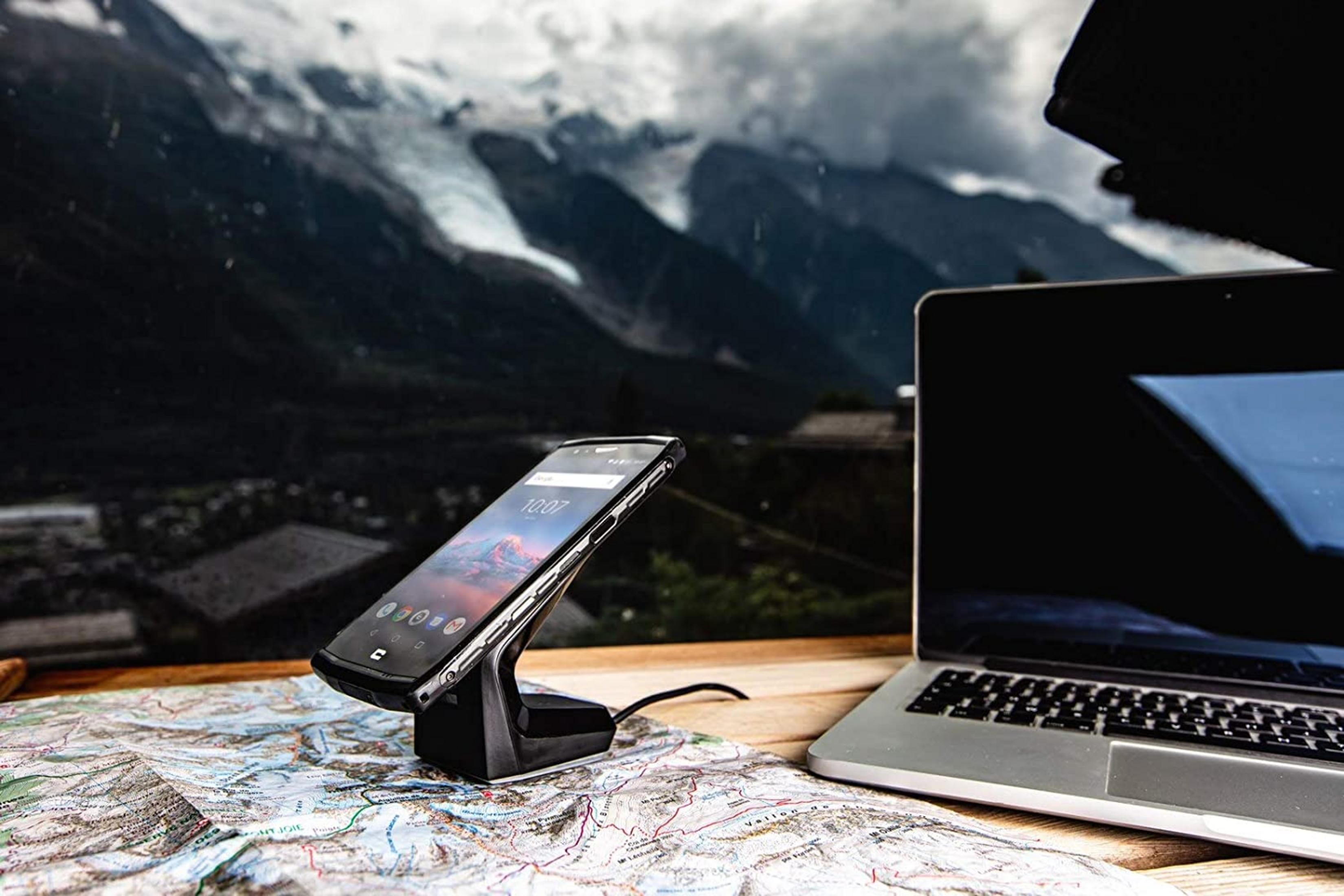Smartphone su supporto, laptop e mappa su tavolo di legno con montagne sullo sfondo.