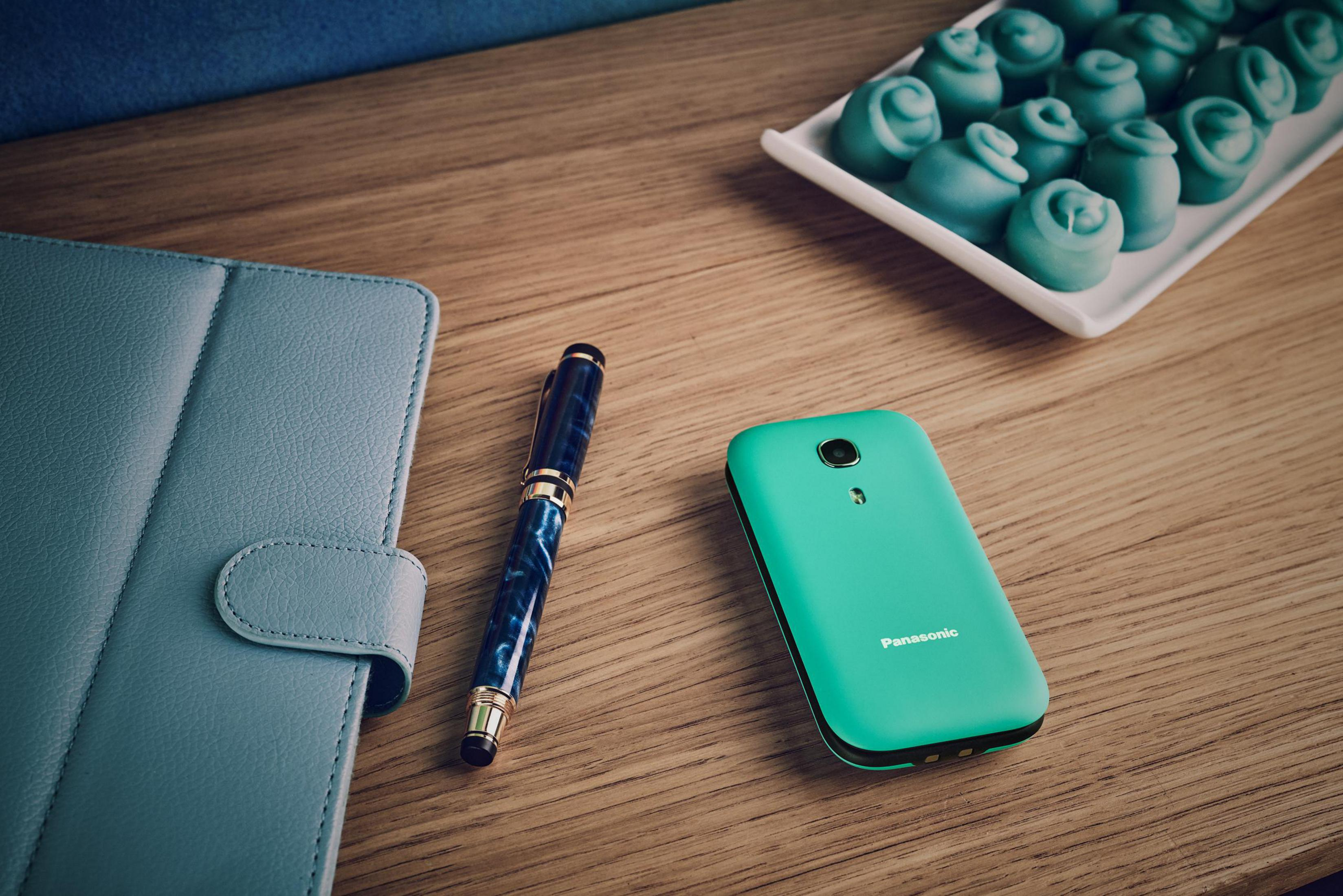 Un telefono Panasonic turchese, una penna stilografica blu e un taccuino poggiano su un tavolo di legno.