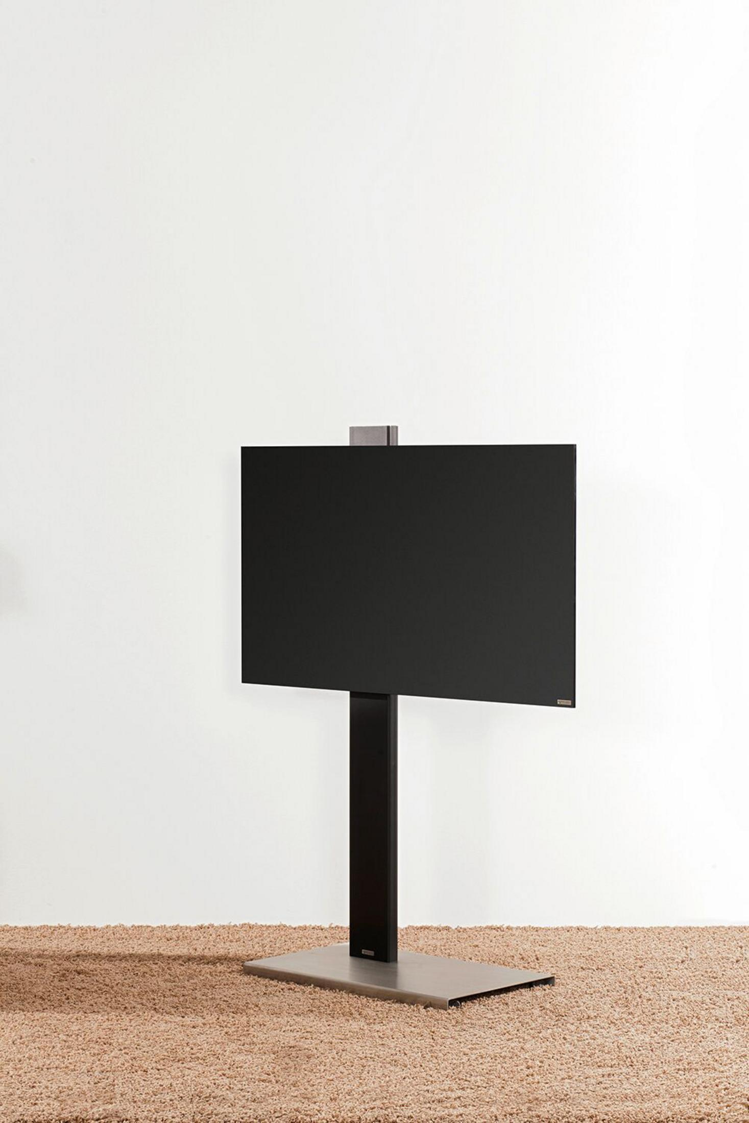 Ein schwarzer Fernseher auf einem dunklen Ständer steht auf einem hellbraunen Teppich vor einer weißen Wand.