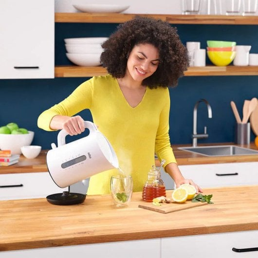 Een vrouw in een gele top schenkt water uit een witte waterkoker in een glas.