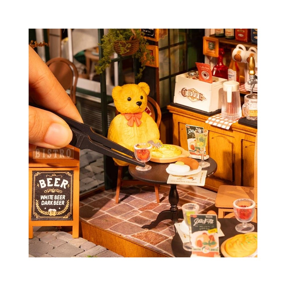 Ein Teddybär sitzt in einem Miniatur-Bistro mit Essen und Getränken auf dem Tisch.