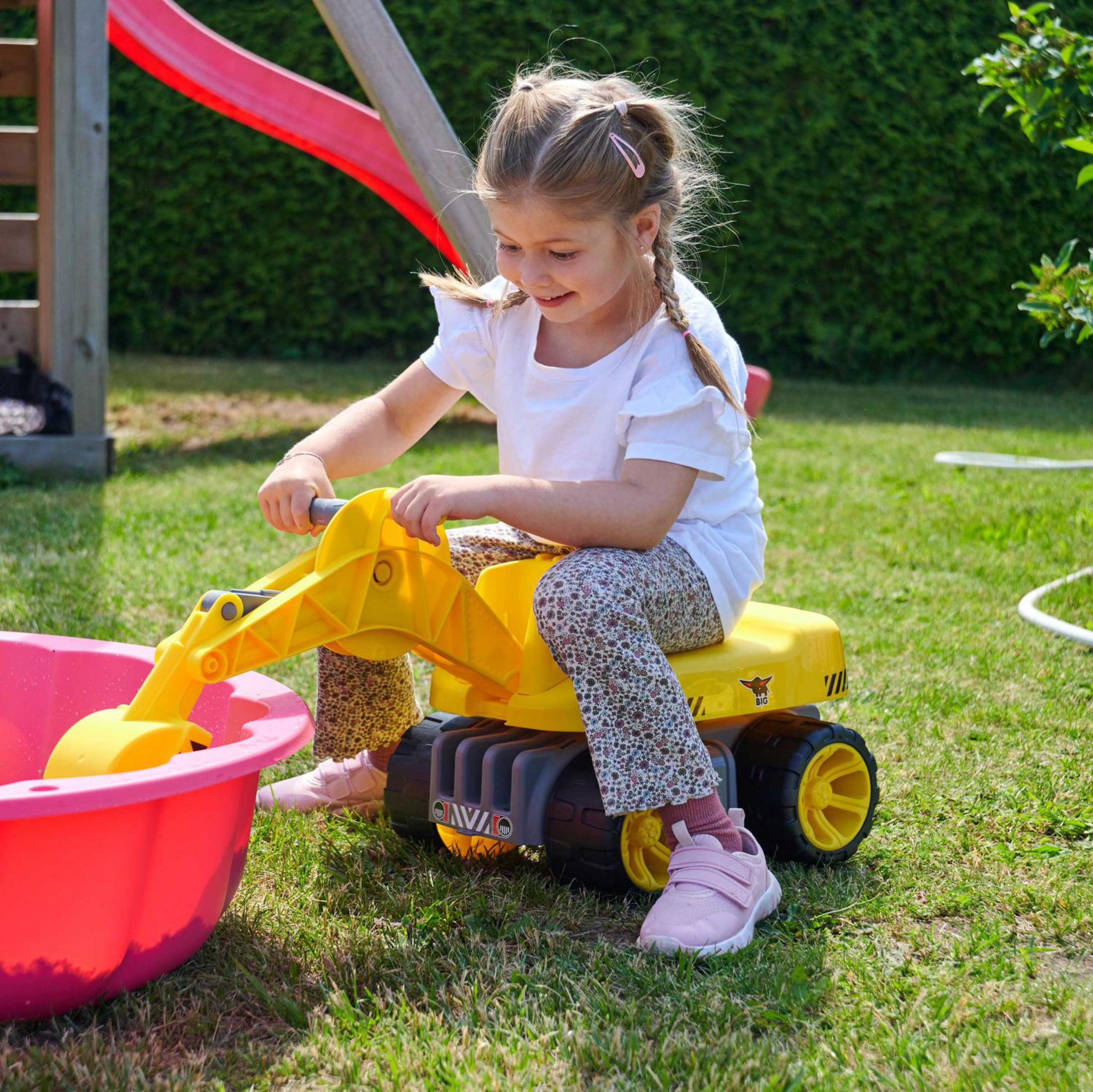 Ein junges Mädchen in weißem Hemd und gemusterter Hose spielt mit einem gelben Spielzeugbagger im Garten.