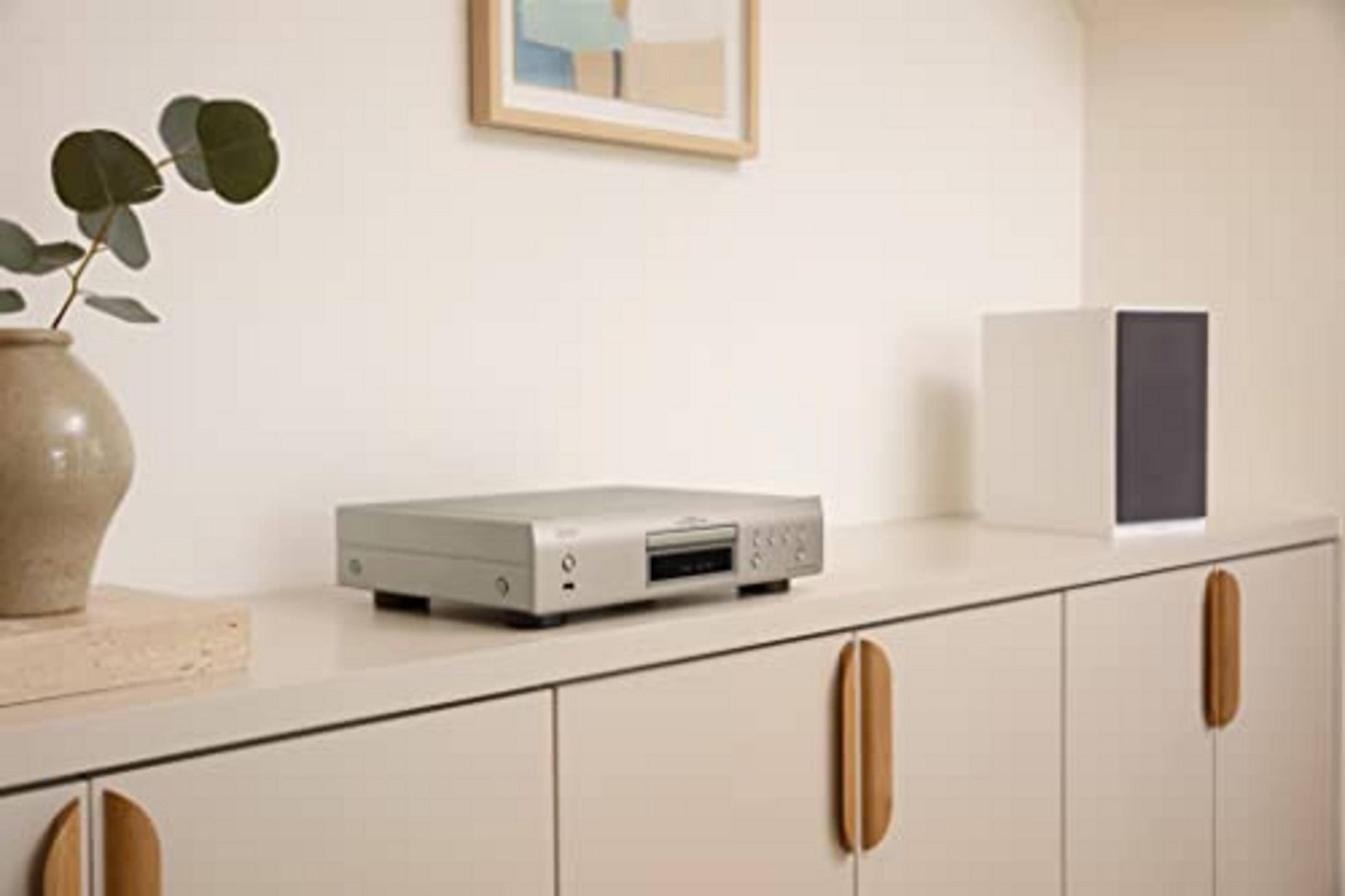 Ein silberner CD-Player steht auf einem hellen Schrank neben einer Vase mit Zweigen.