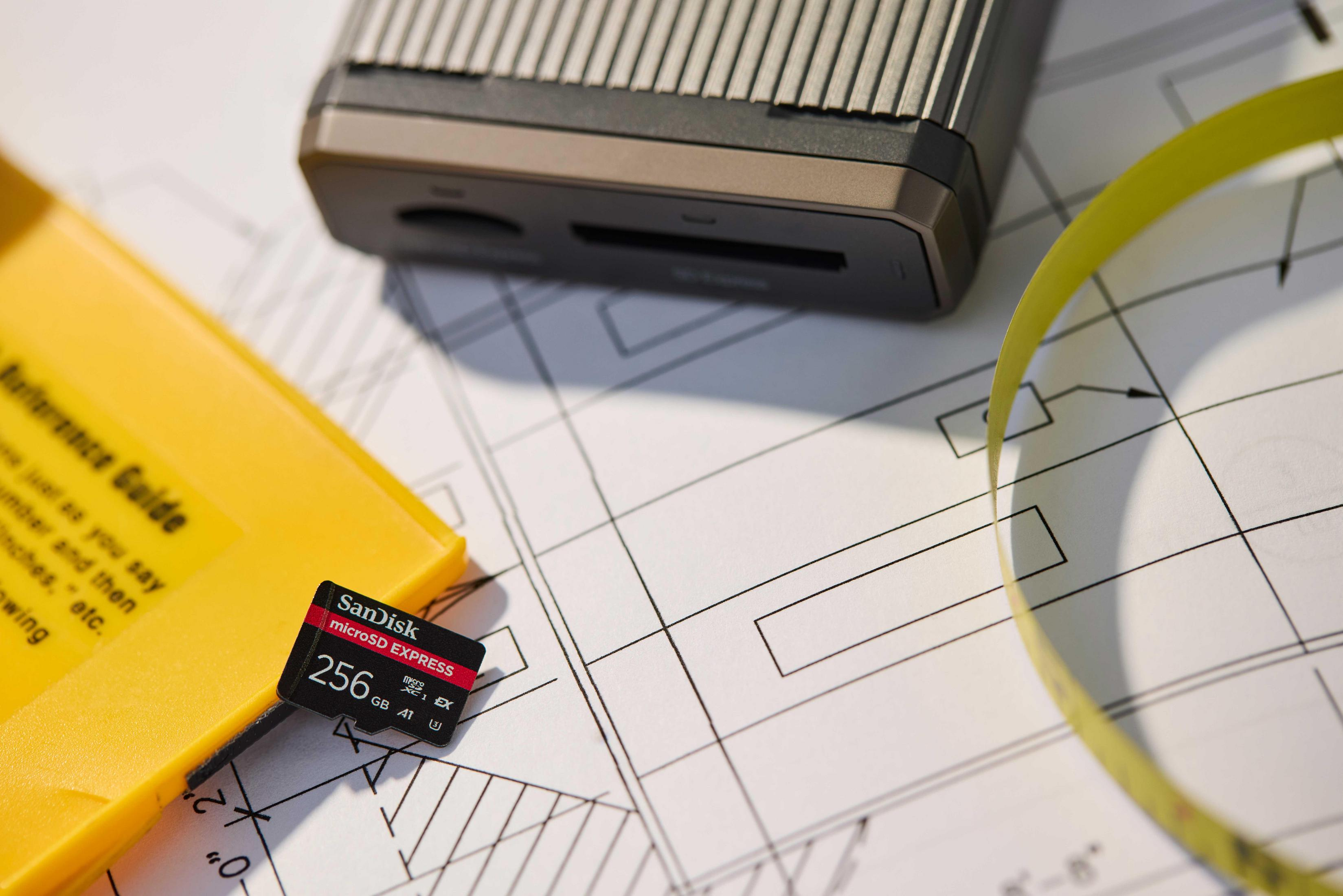 Una tarjeta microSD SanDisk sobre planos junto a una caja amarilla y cinta métrica.