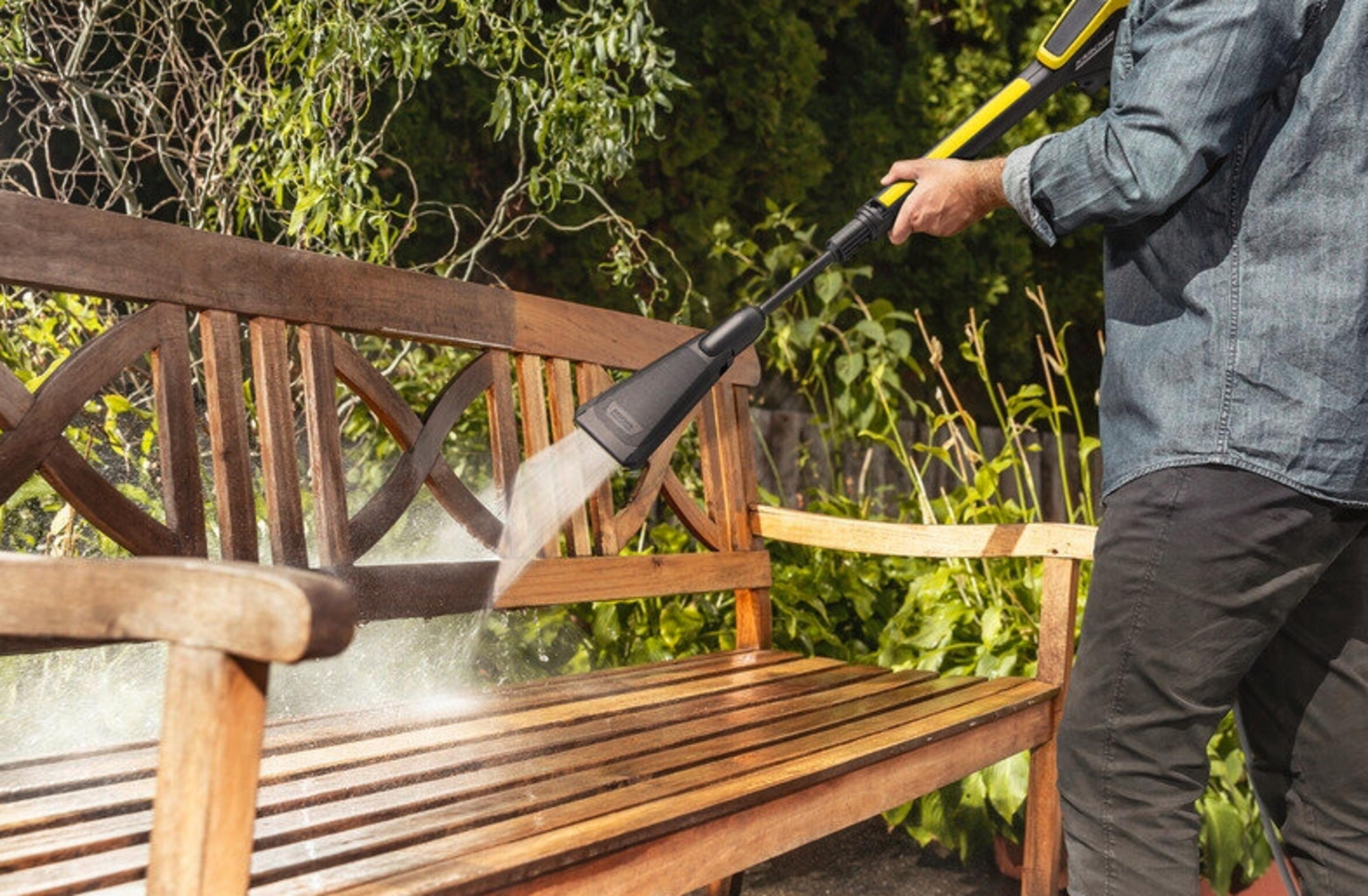 Un uomo pulisce una panchina di legno con un'idropulitrice in un giardino.