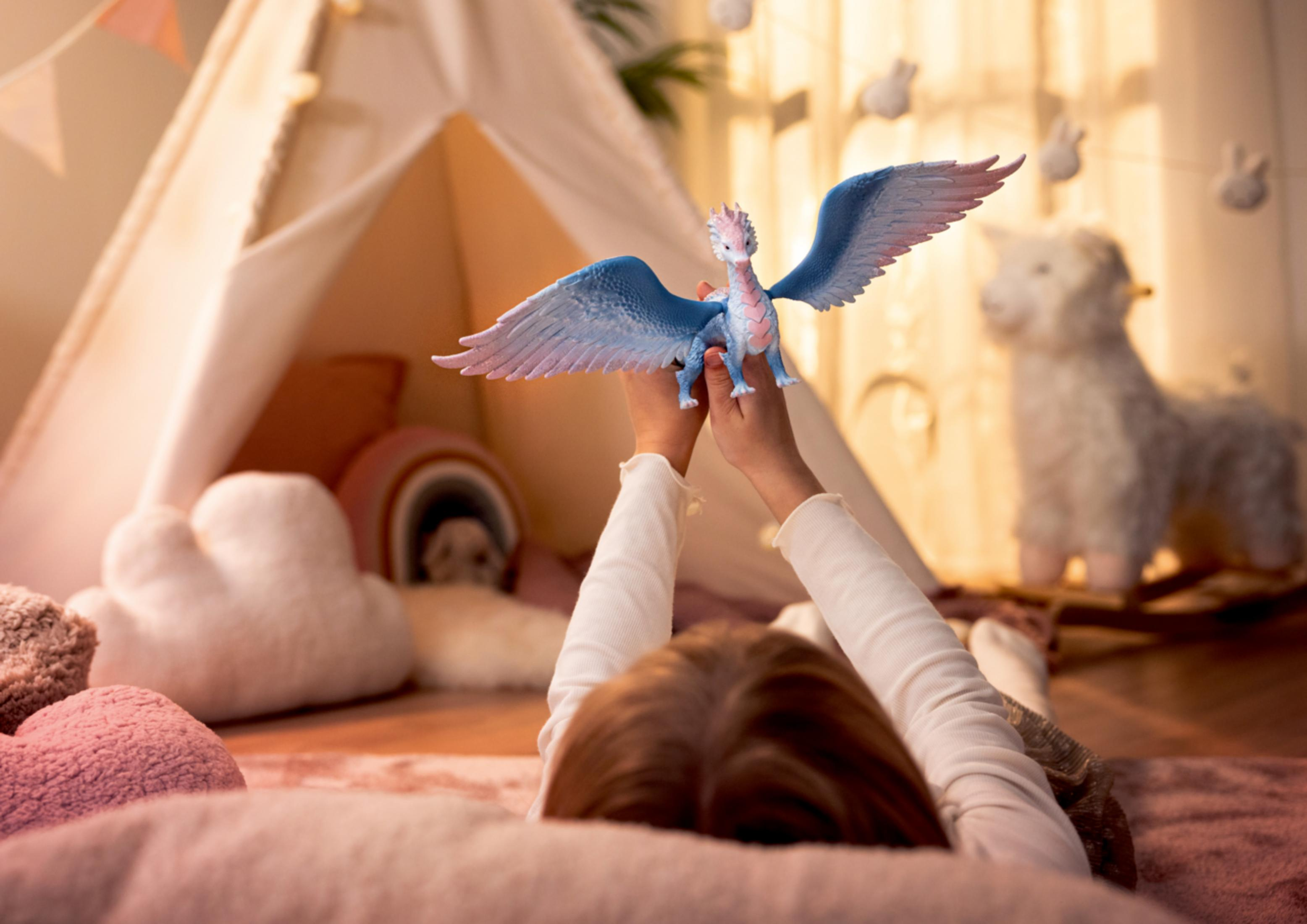 Un bambino tiene un giocattolo drago blu e rosa davanti a una tenda da gioco con ghirlanda di conigli.