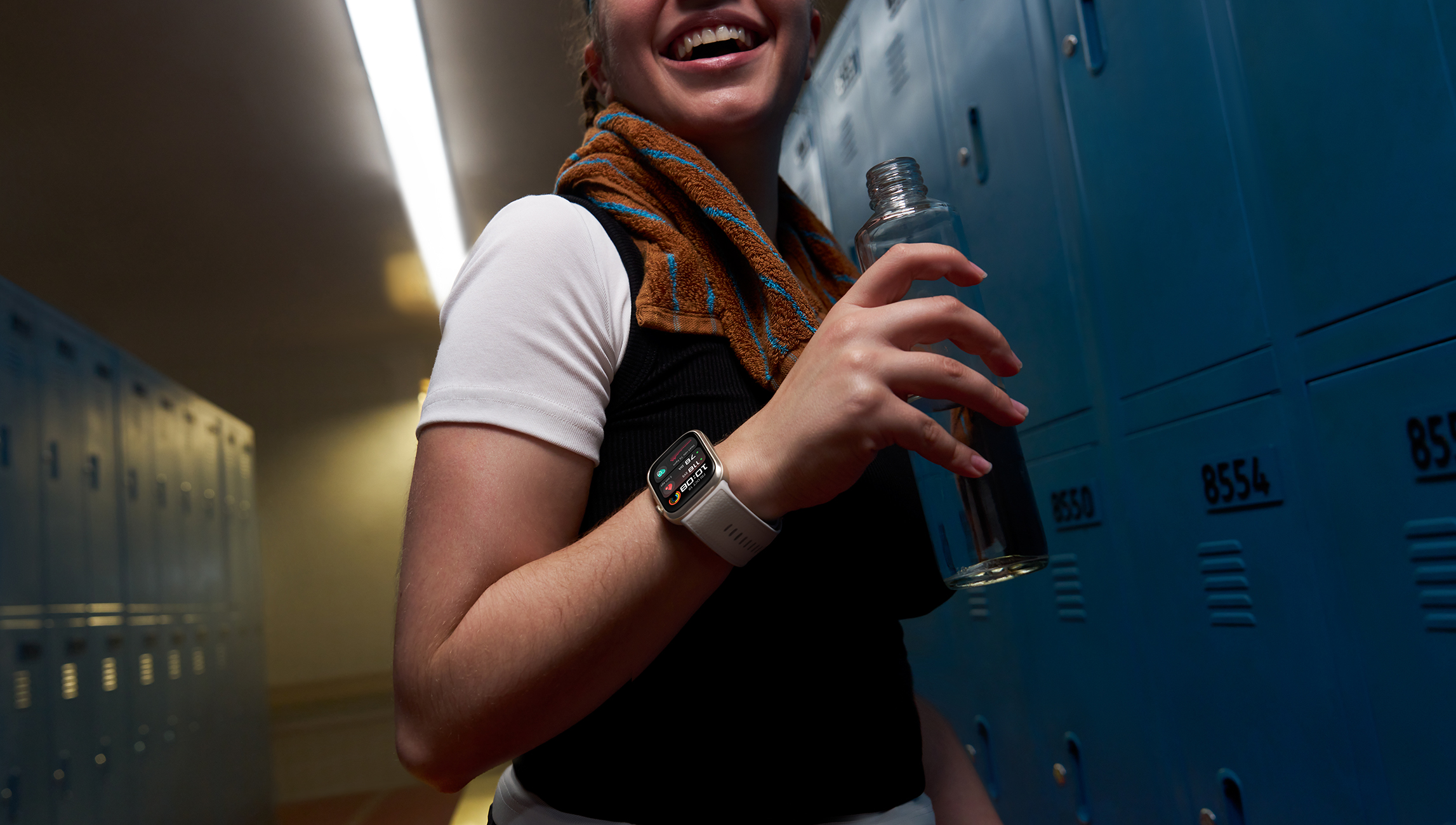 Eine lachende Person in Sportkleidung hält eine Wasserflasche neben blauen Schließfächern.