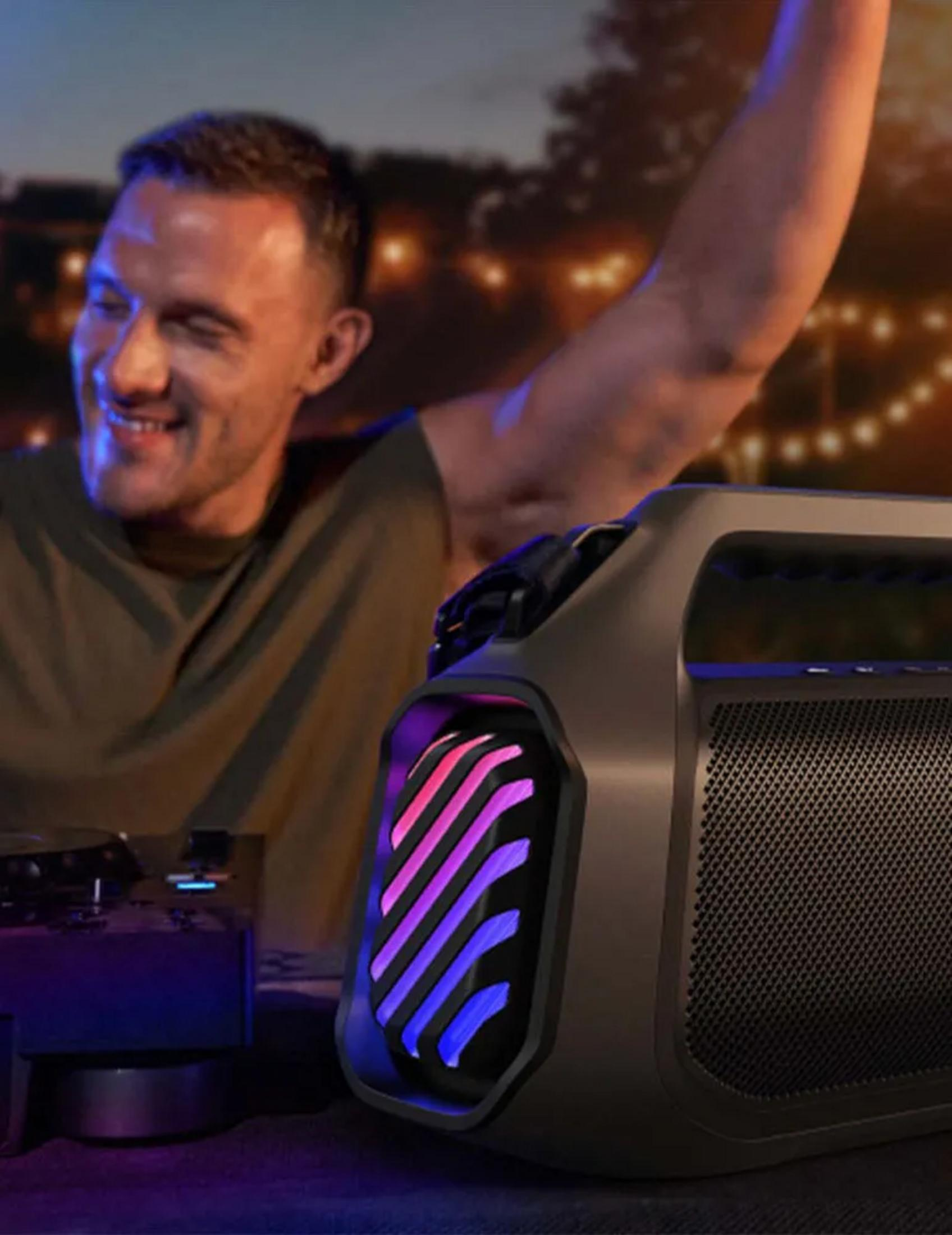 Uomo con braccio alzato balla vicino a speaker portatile con luci colorate e DJ set.