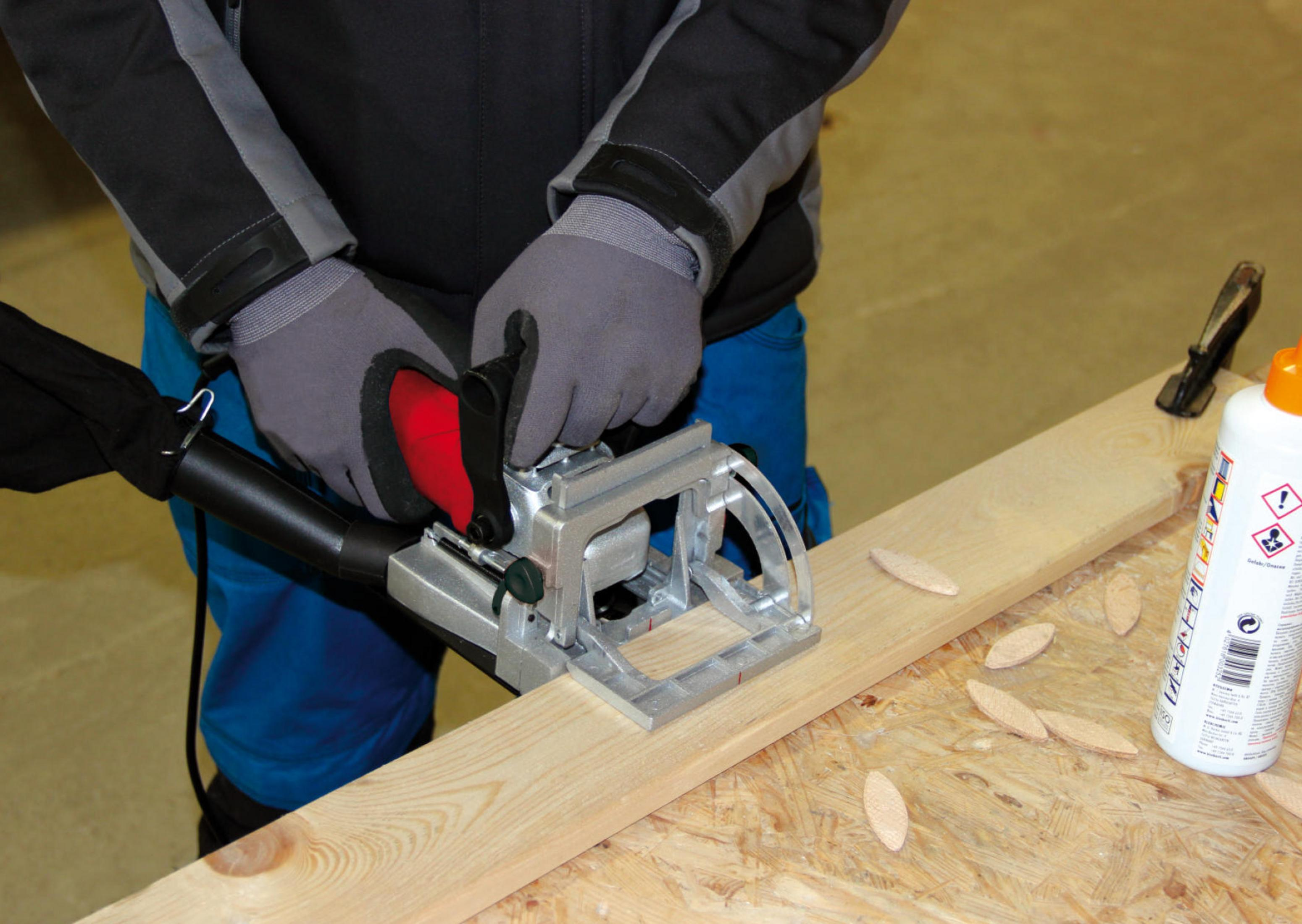 Mann mit Handschuhen benutzt Dübelmaschine auf Holzbrett. Kleine Holzdübel liegen daneben.