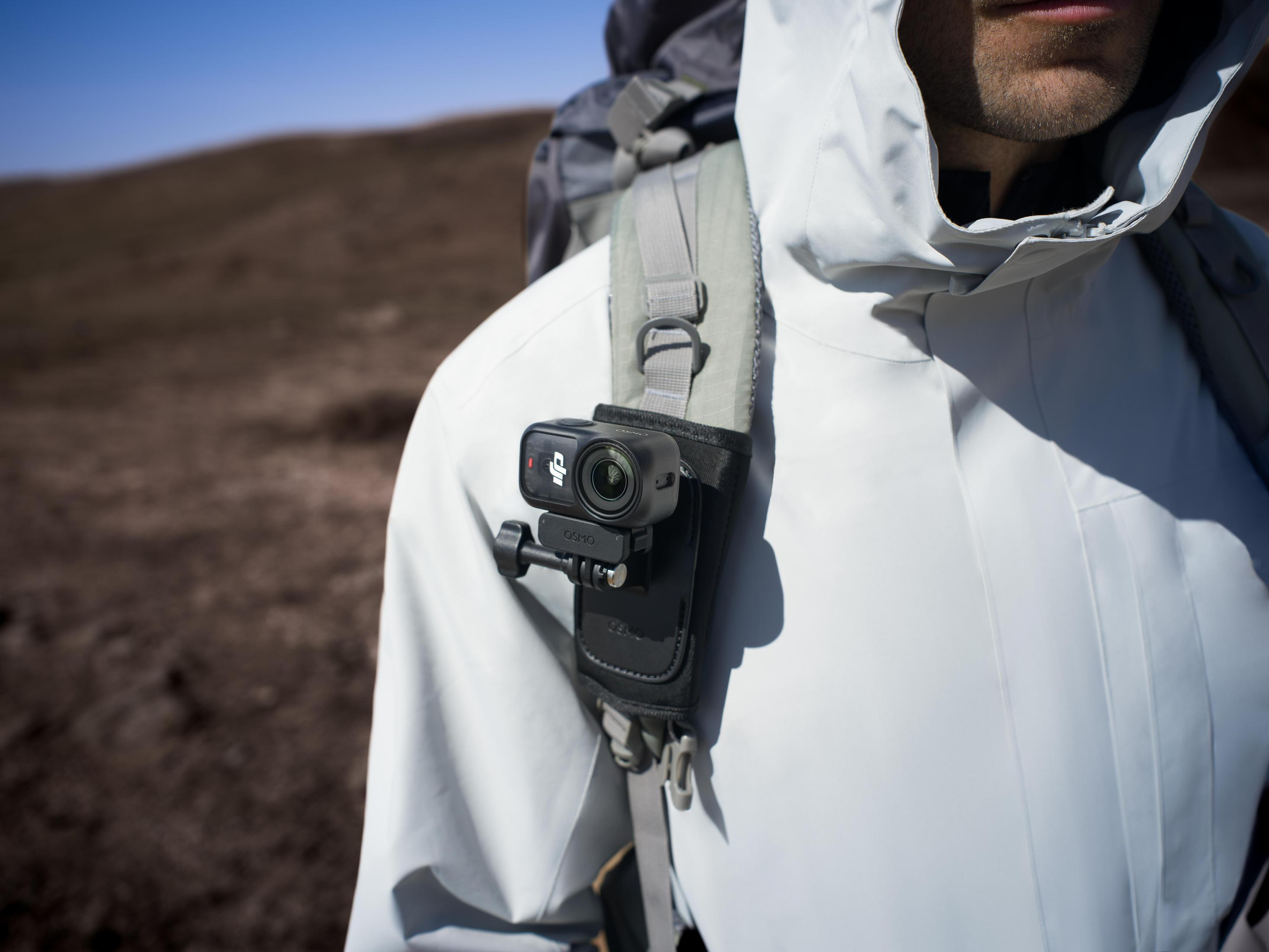 Een man draagt een witte jas en rugzak met een camera op zijn borst.