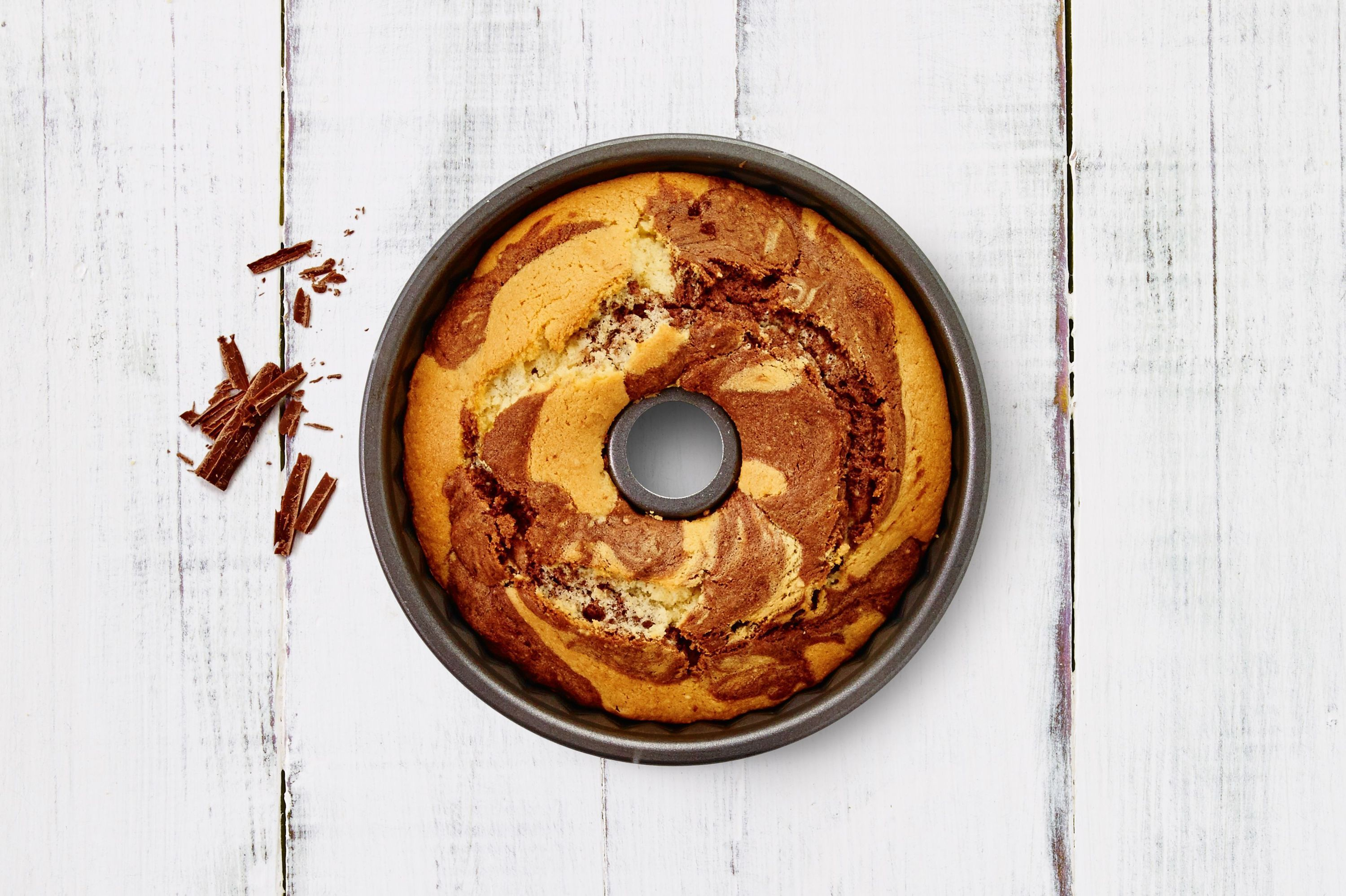 Marmorkuchen in einer Gugelhupfform auf weißem Holz, daneben Schokostückchen.