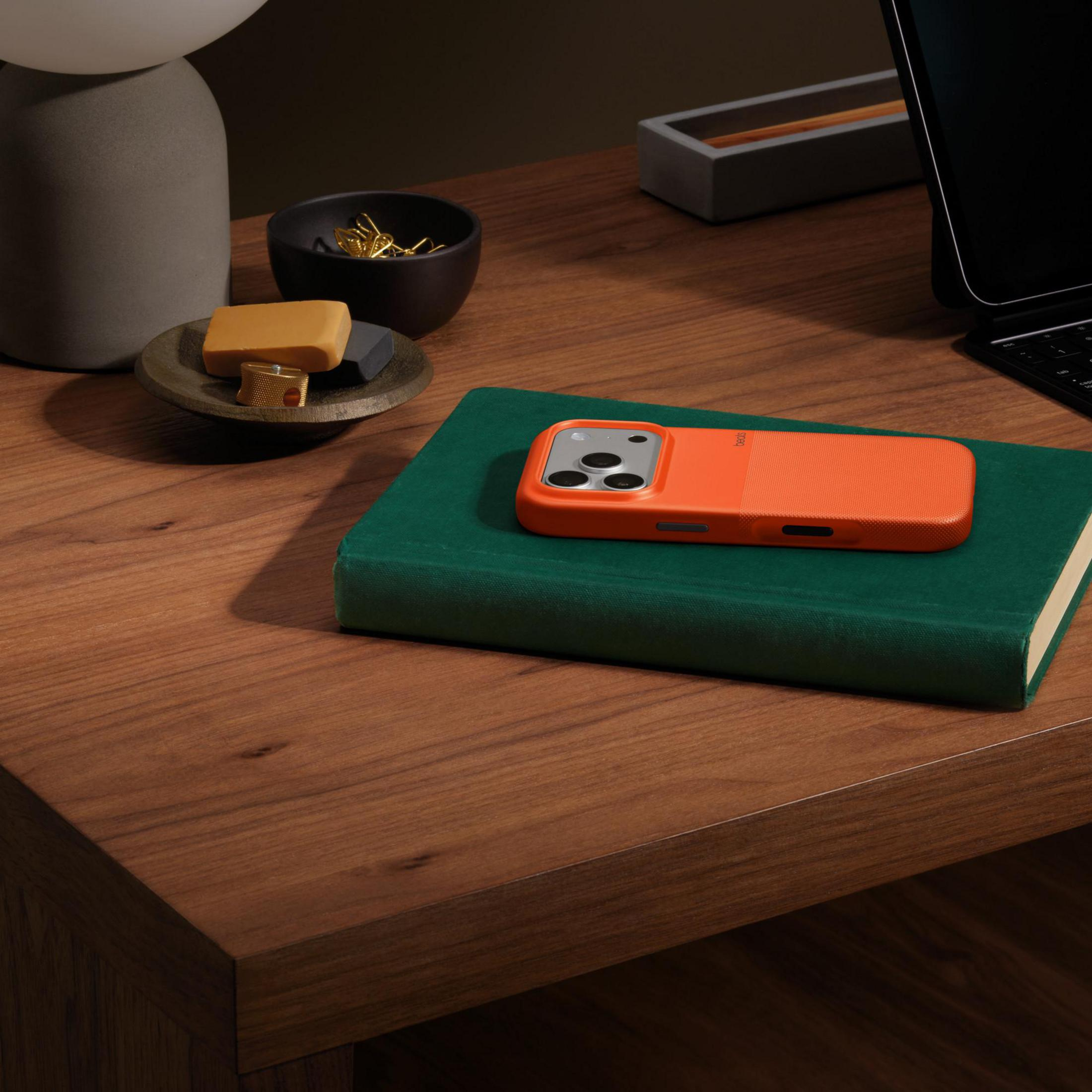 Ein oranges Handycase liegt auf einem grünen Buch, daneben Stifte und eine Lampe.