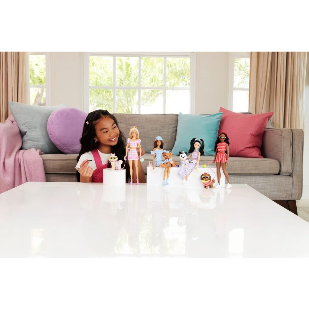 Una niña juega con muñecas Barbie y mascotas en una mesa blanca frente a un sofá.