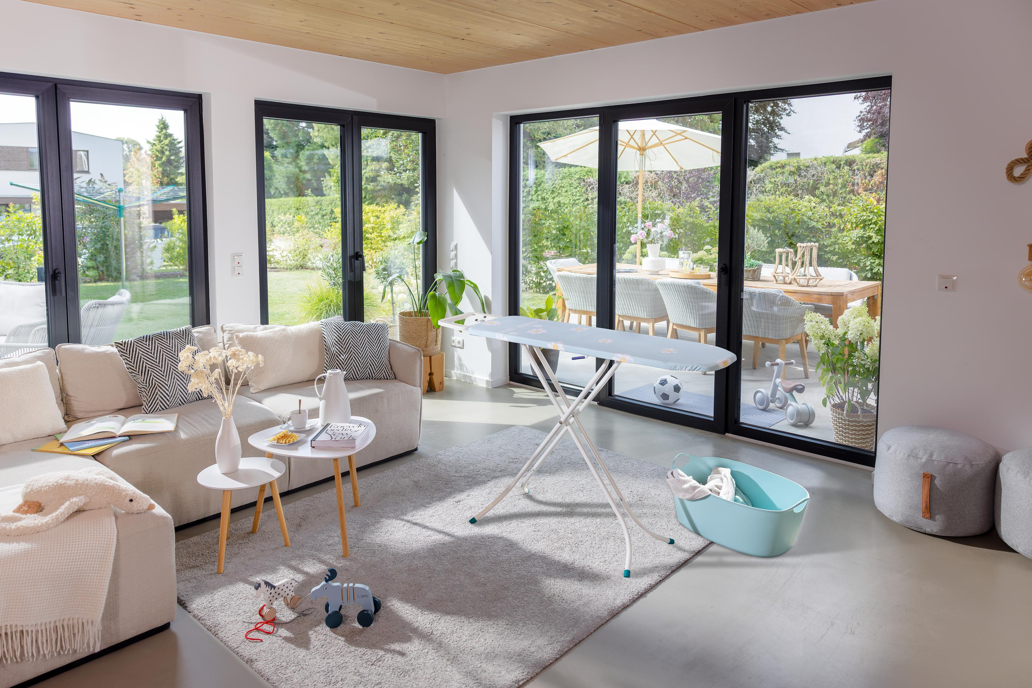 Ein Wohnzimmer mit Sofa, Büchern, Spielzeug und einem Bügelbrett, das zum Garten führt.
