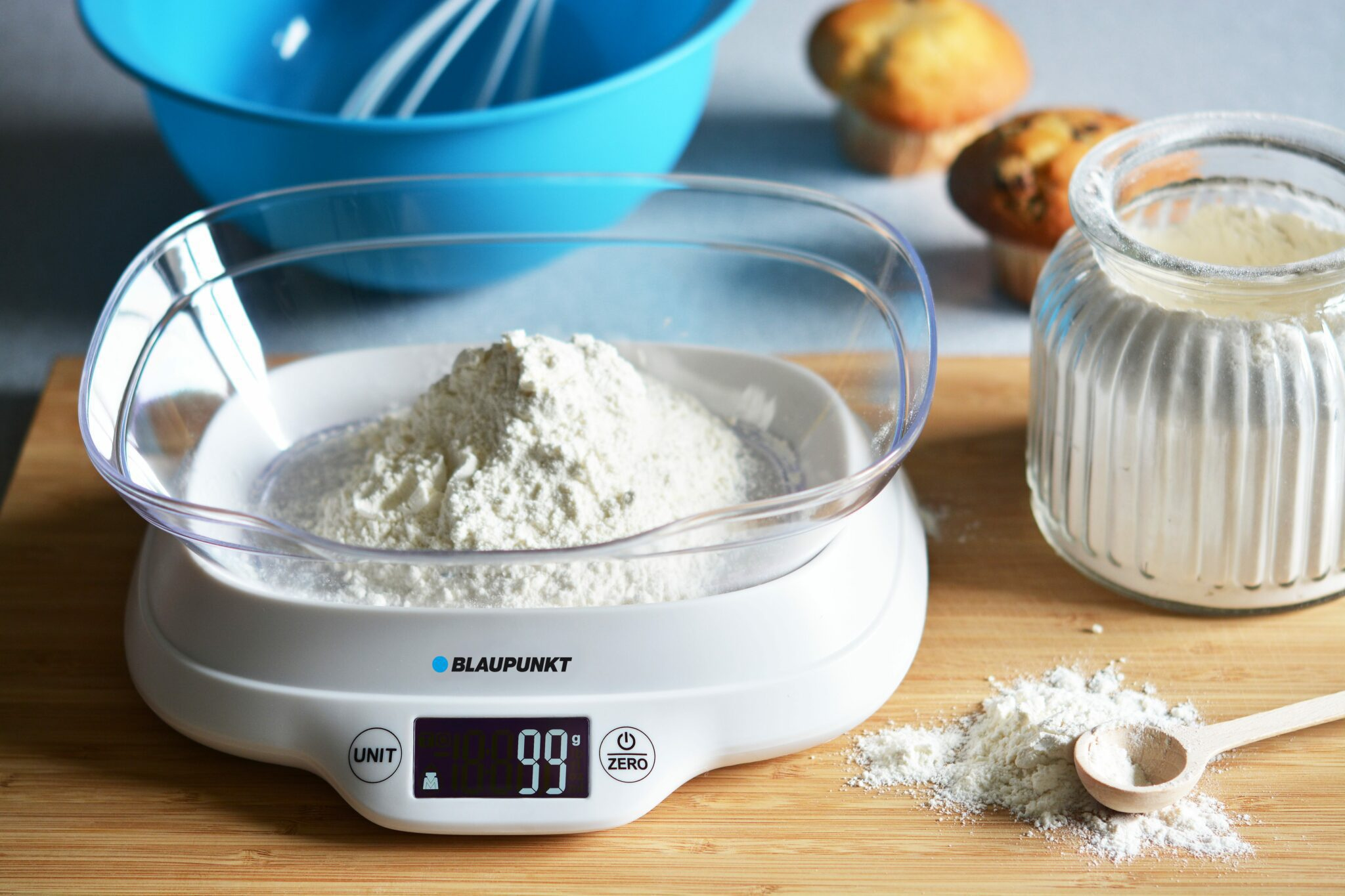 Una báscula digital muestra 99g de harina. Un bol azul y muffins están al fondo.