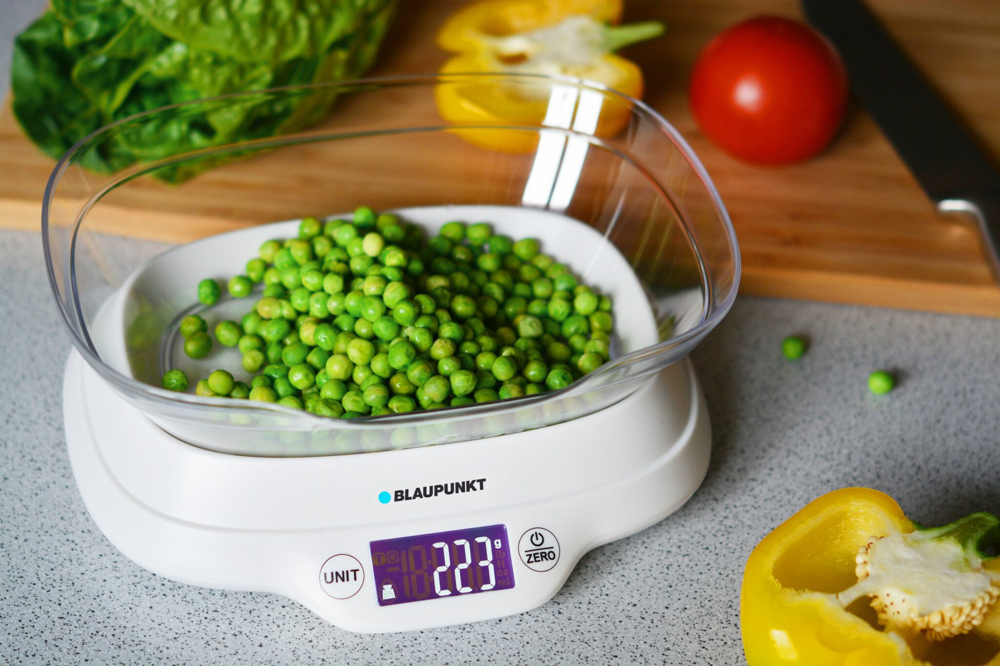 Una báscula de cocina digital con guisantes, lechuga, tomate y pimiento.