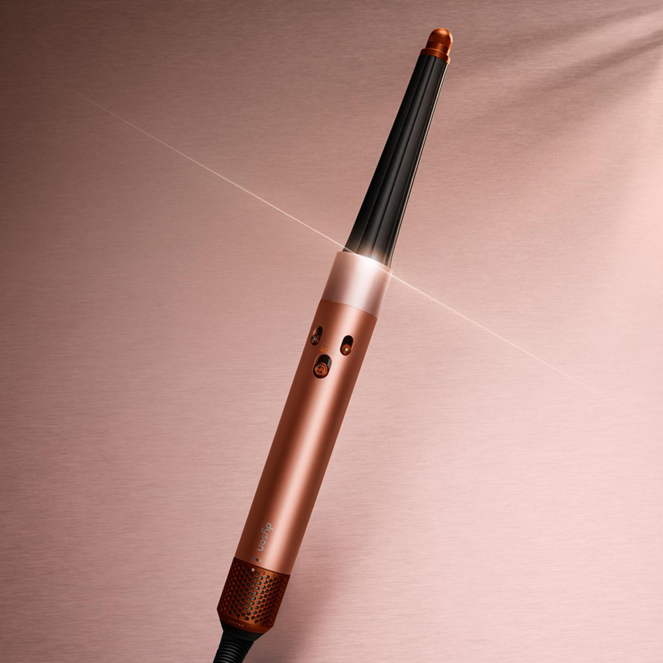 Un styler per capelli Dyson color bronzo con cilindro nero su sfondo testurizzato oro rosa.